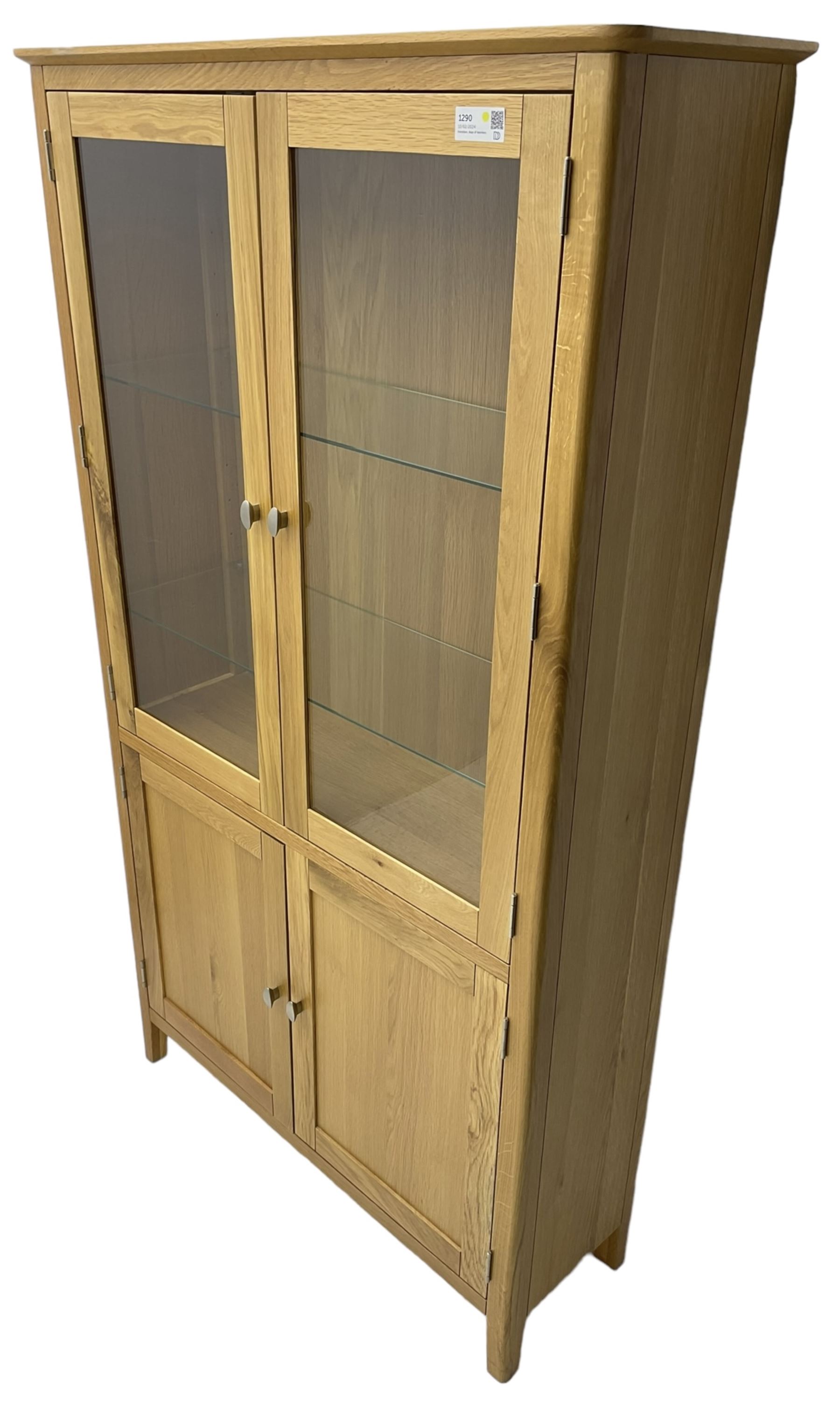 Contemporary light oak display cabinet on cupboard, fitted with two glazed doors enclosing two adjustable glass shelves with light fittings to the top, over panelled cupboard concealing single shelf, on square supports