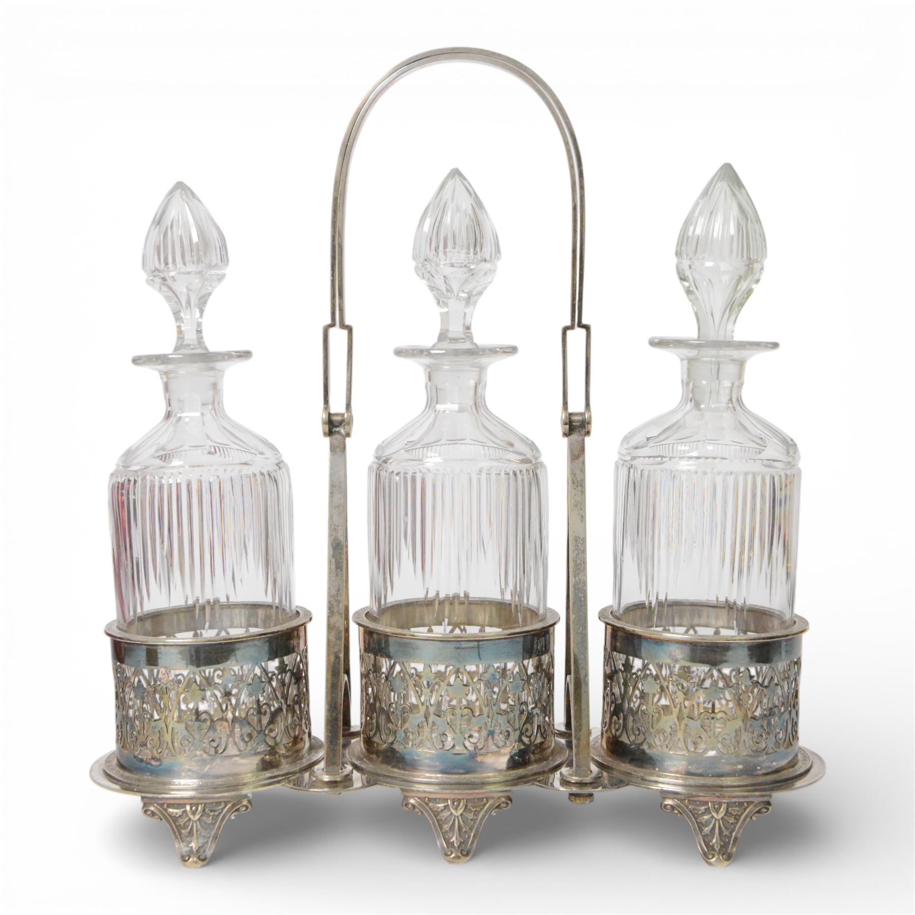 Late 19th century silver plated Silber & Fleming triple decanter stand, the three cut glass decanters with spire stoppers housed within foliate and scroll pierced bottle sleeves, upon a tripart base with six scroll feet, the base mounted with articulated x-frame curved handle, including handle when positioned upright H35.5cm, W34cm