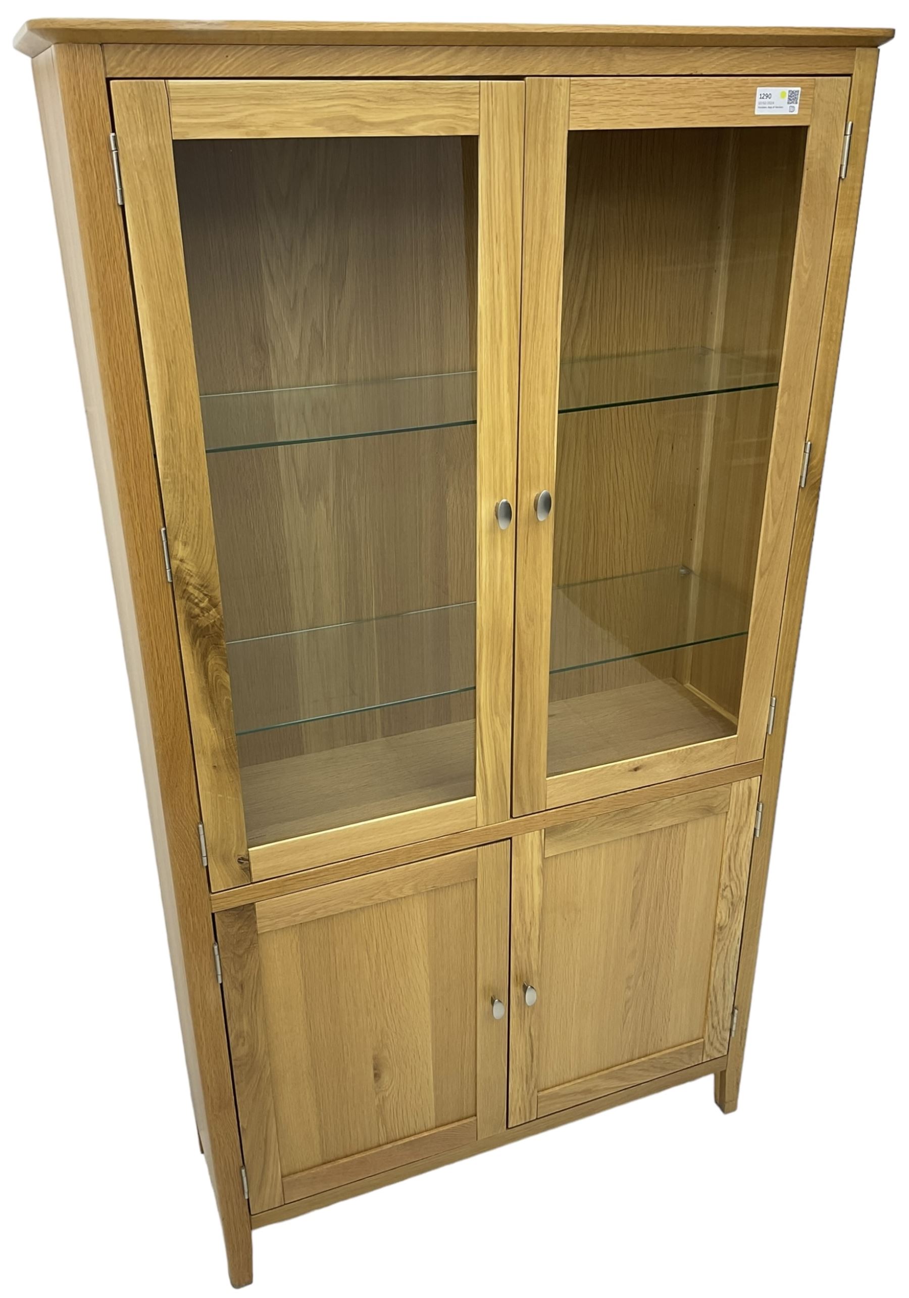 Contemporary light oak display cabinet on cupboard, fitted with two glazed doors enclosing two adjustable glass shelves with light fittings to the top, over panelled cupboard concealing single shelf, on square supports