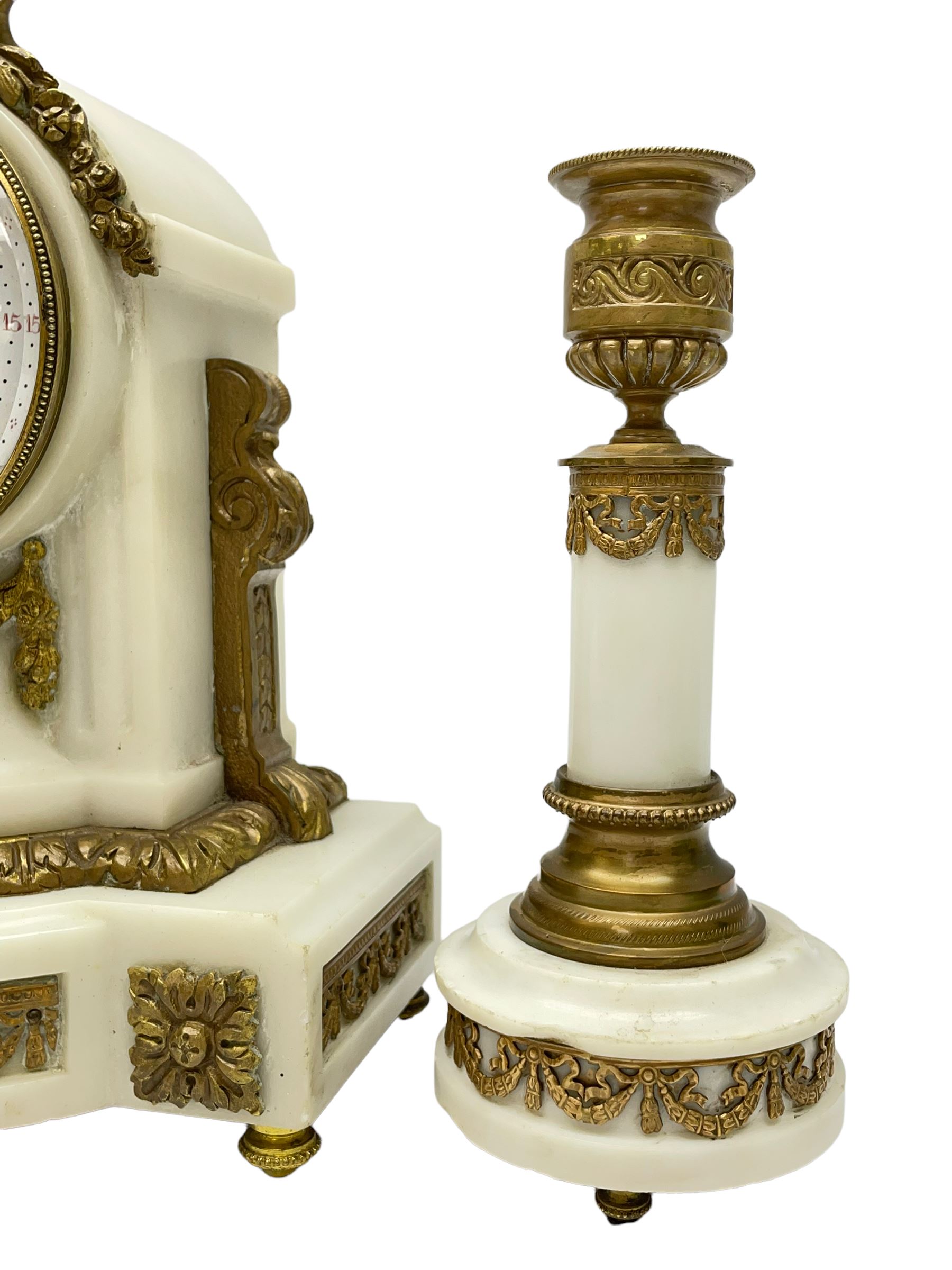 An early 20th century white marble mantle clock in a break front case with an arched top and finial, gilded scroll work to the sides and pierced foliate panels to the front, plinth raised on five bun feet, enamel dial with garland decoration, Arabic numerals, minute markers and Louis XV gilt hands, within a convex glazed bezel, eight-day Parisian striking movement striking the hours and half hours on a bell, with two conforming single light candlesticks. With Pendulum.

