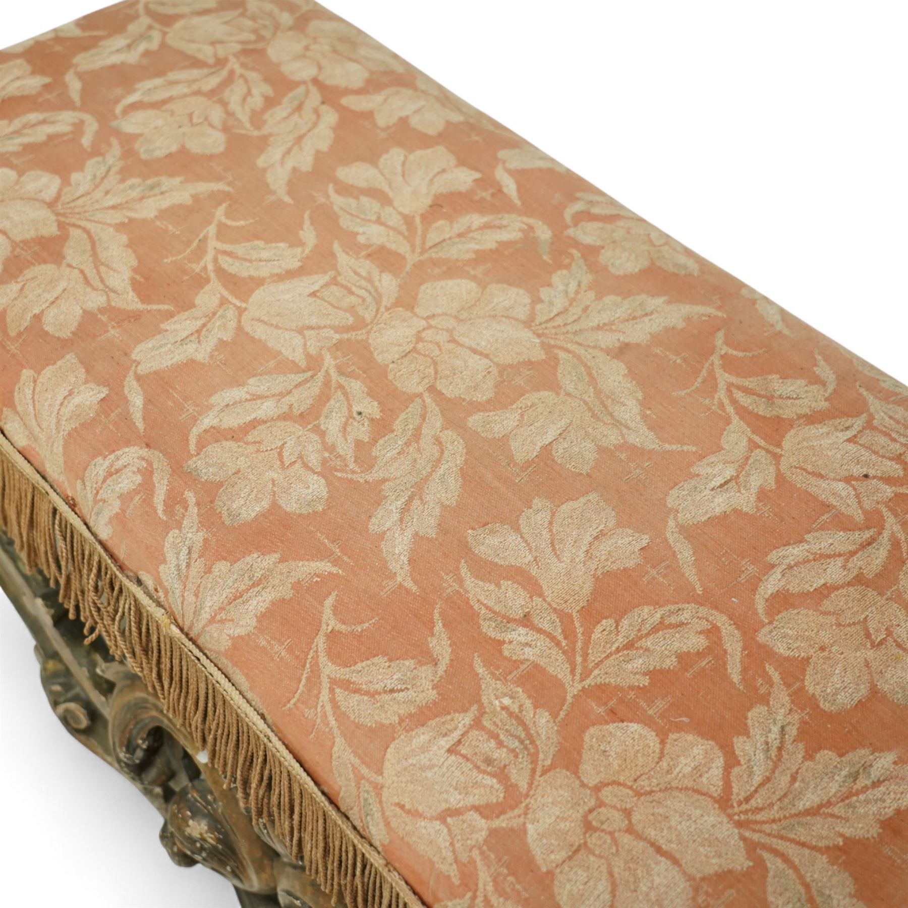 19th-century Rococo-style giltwood window seat, upholstered in floral-patterned coral-ground fabric with fringed border, the intricate scrolled supports united by pierced scrolling foliate stretchers with acanthus and C-scroll decoration