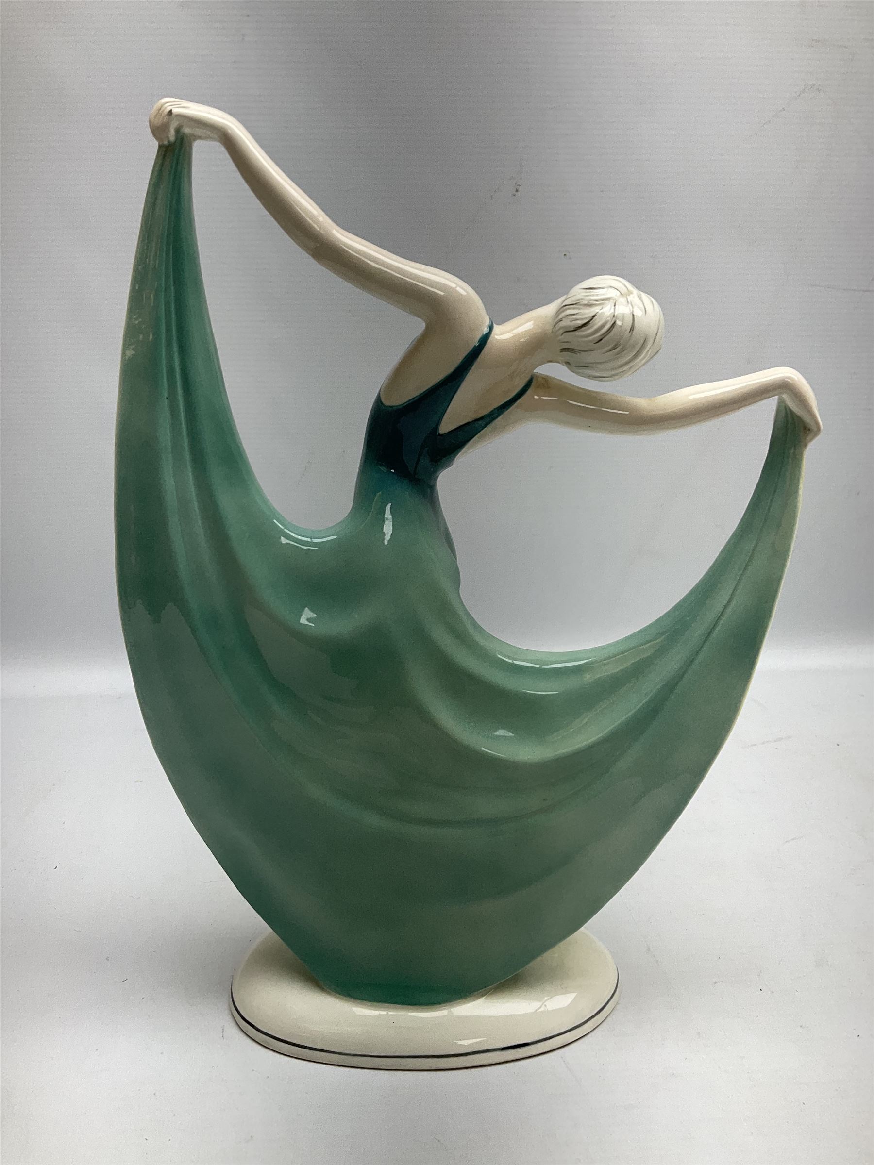 Early 20th century Hertwig Katzhütte figure of an Art Deco lady dancing, wearing a green, blue and yellow dress, stood upon on a domed plinth, with green cat H house printed mark beneath, H32cm