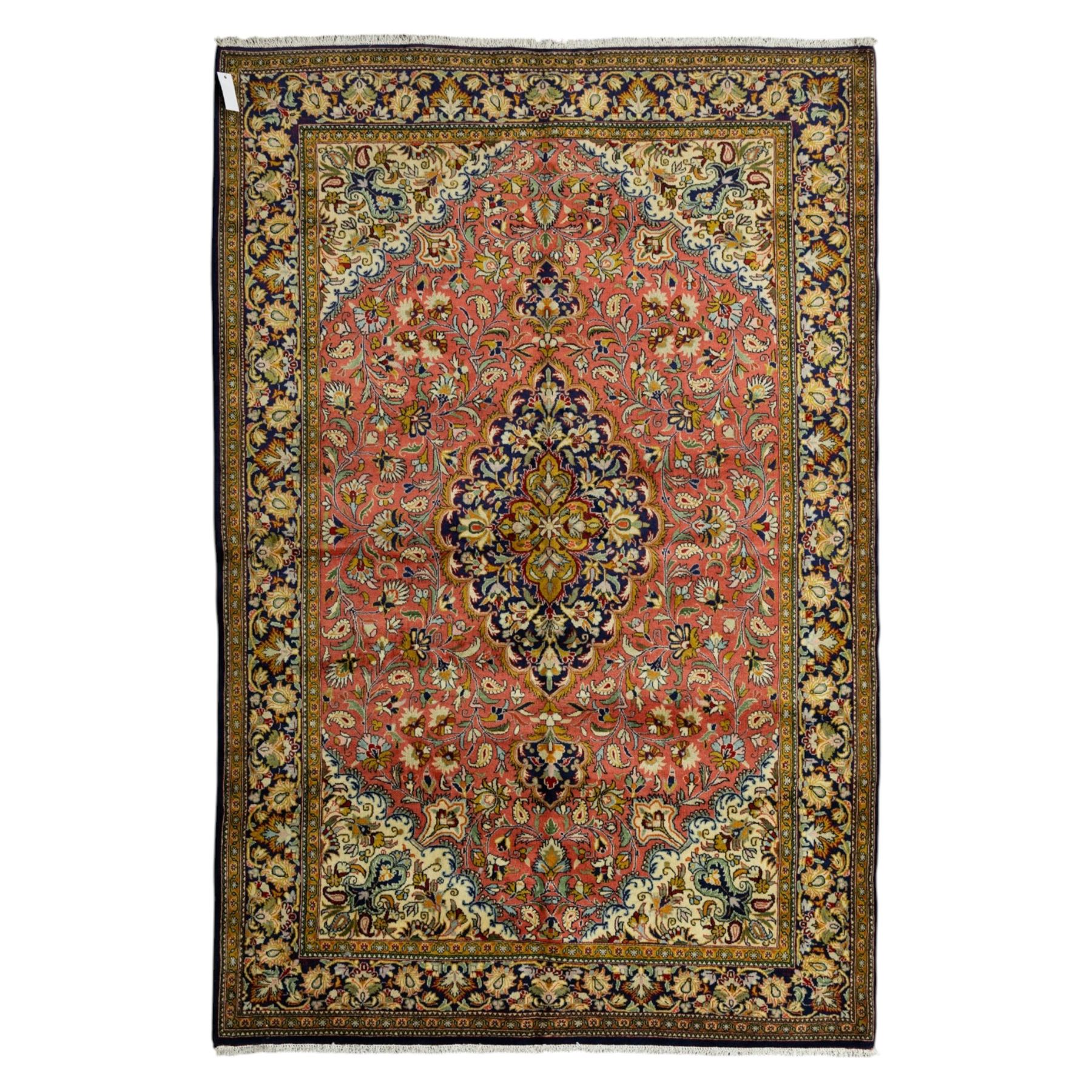 Central Persian Qom hand-knotted carpet, salmon pink ground, with a central dark navy shaped floral medallion with pendants, surrounded by dense scrolling foliate and floral motifs incorporating rosettes, palmettes and delicate vine work, enclosed by ivory spandrels and a dark navy border with repeating palmettes and meandering vines, flanked by narrow floral guard stripes, with cotton fringes