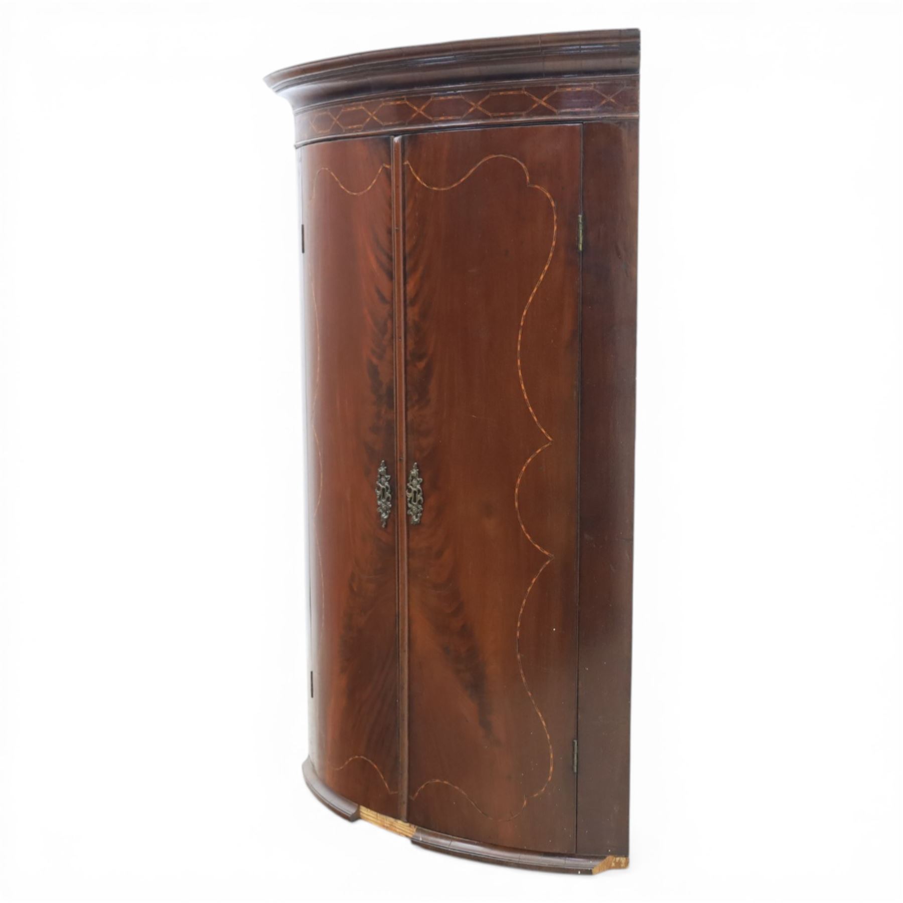 George III mahogany bow-front corner cupboard, projecting moulded cornice over satinwood inlaid frieze with geometric crossbanding, enclosed by twin shaped panel doors with line inlay and ornate brass escutcheon plates, painted interior fitted with shaped shelves, on moulded plinth base