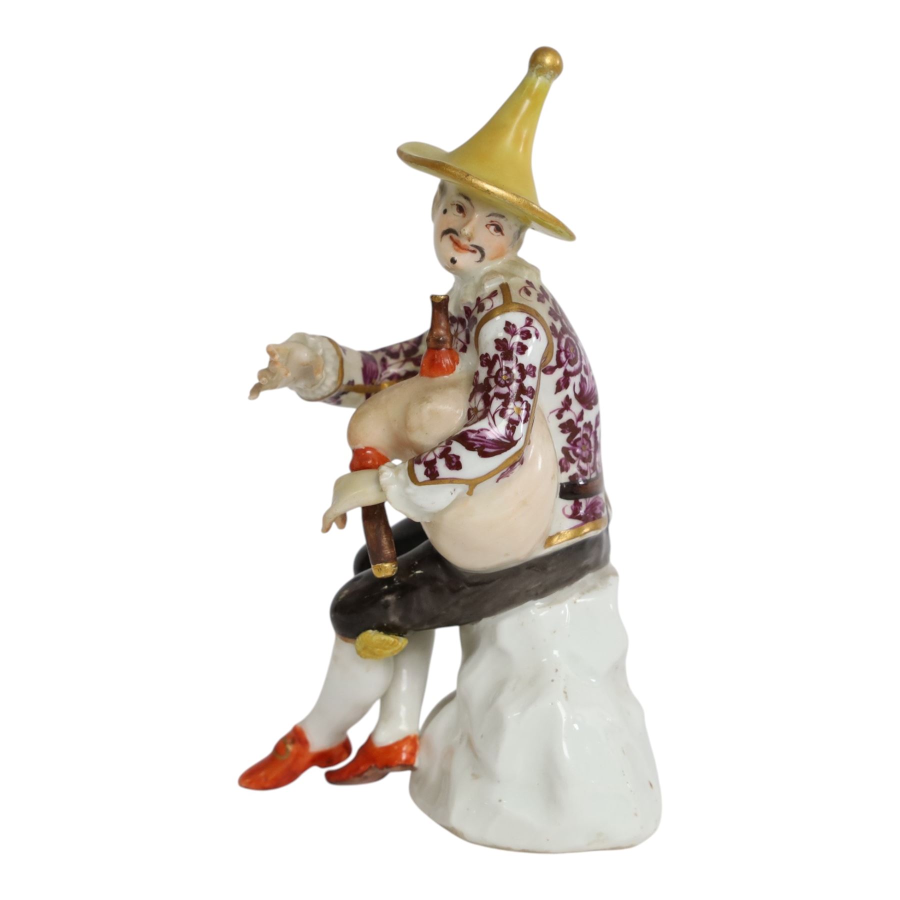 Mid 18th century Meissen figure, modelled as a harlequin playing bagpipes, circa 1745, possibly modelled by J.F.Eberlein after earlier models by J.J.Kändler, wearing a gilt edged tunic painted with flowers, black breeches with rosettes at the knees, red shoes and conical yellow hat, seated upon a rock, with traces of blue crossed swords mark beneath, H14cm