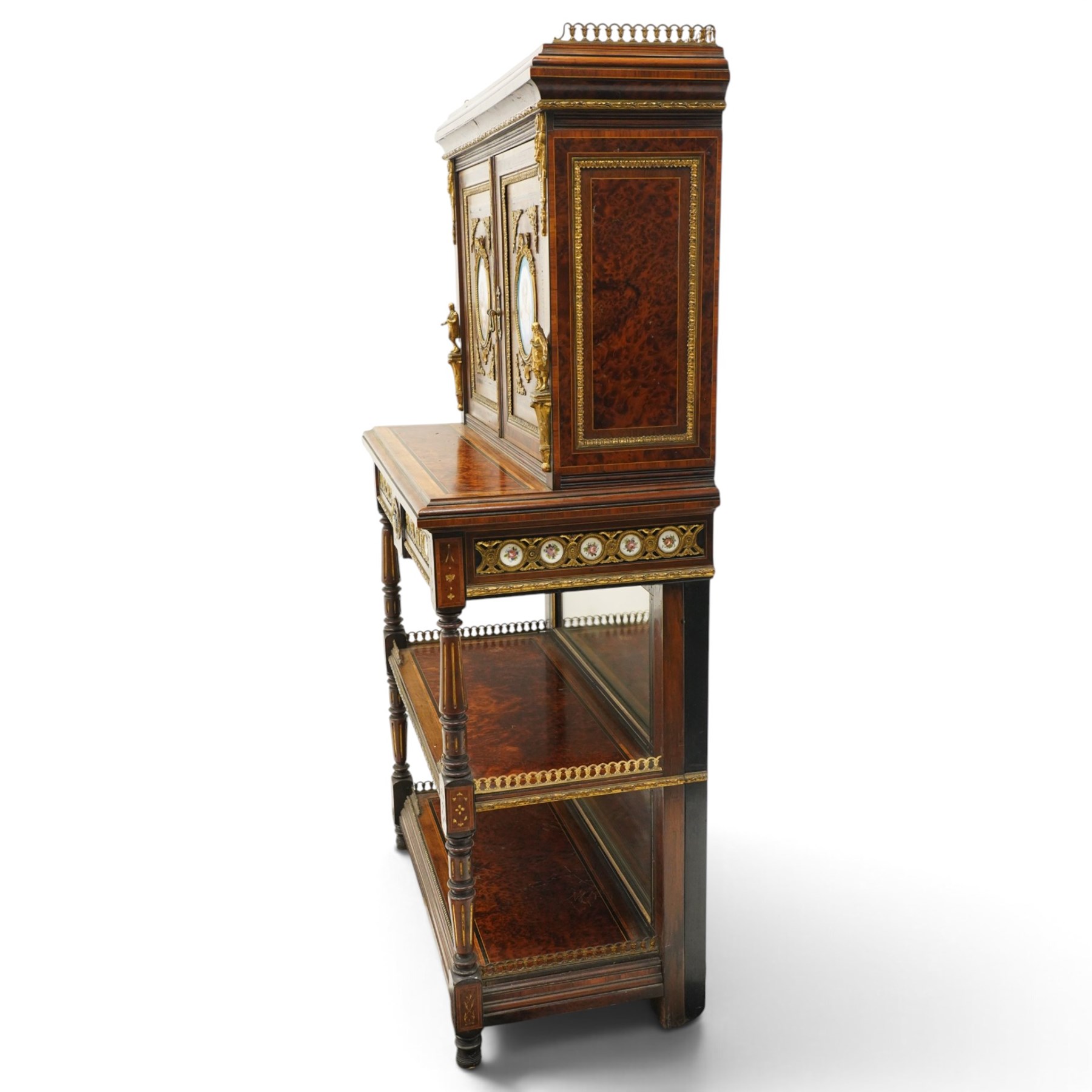 Fine late 19th century Aesthetic Movement ebony and amboyna cabinet on stand with parcel-gilt detail, the moulded cornice with pierced gilt-brass three-quarter gallery above a pair of panelled doors centred by oval porcelain portrait plaques within gilt-metal ribbon-tied frames, the doors further enriched with gilt-metal foliate mounts and flanked by classical figural terms, enclosing a shelved interior, the projecting lower section with amboyna top above a frieze drawer set with a series of floral porcelain roundels within pierced gilt-metal mounts and a central oval floral plaque, raised on turned and fluted supports with gilt-heightened detail, the mirrored back behind two amboyna shelves with pierced gilt-brass galleries, on turned feet