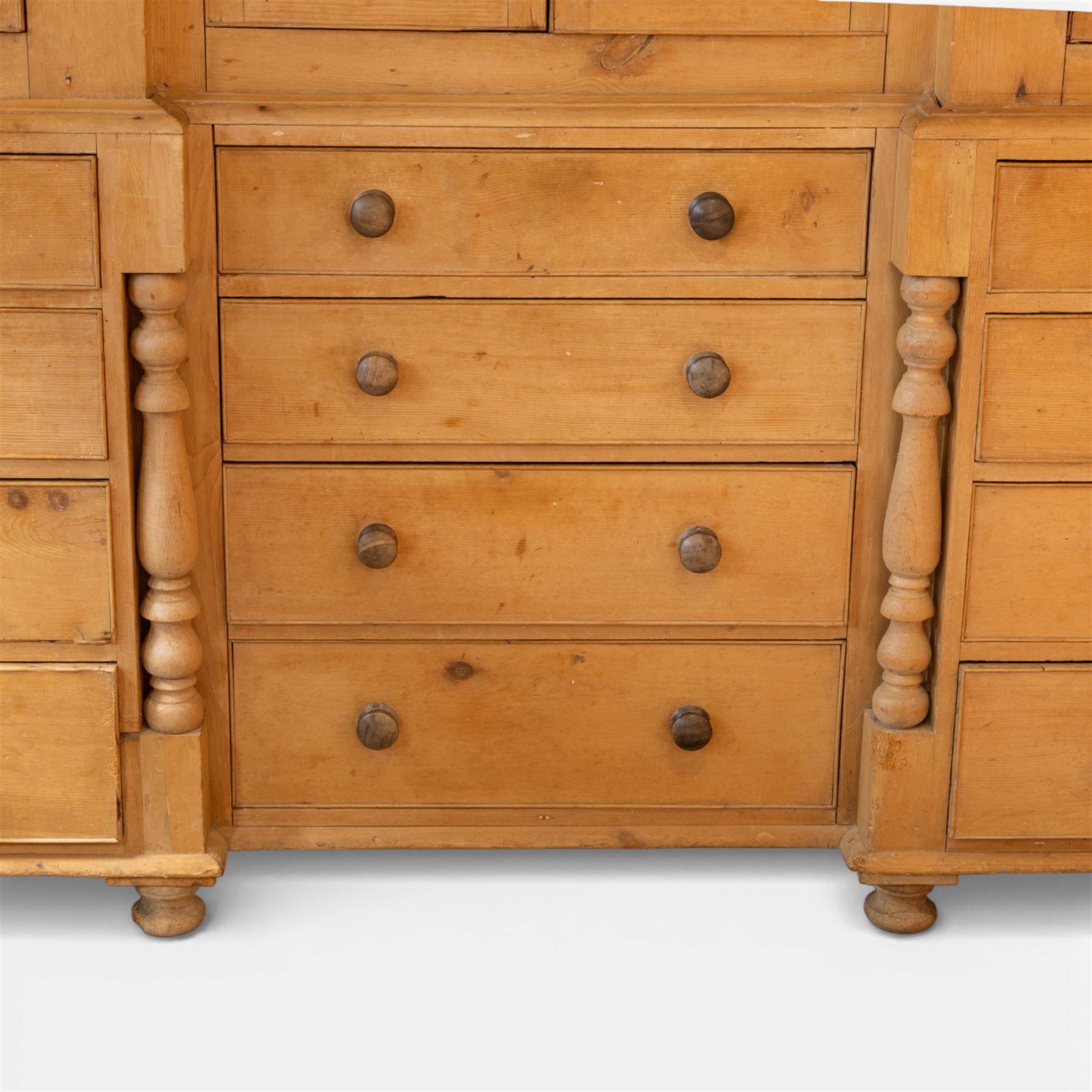 Large Victorian stripped pine reverse breakfront housekeeper’s cupboard, the moulded cornice above a central pair of panelled cupboard doors flanked by single cupboard doors and divided by turned baluster pilasters, the lower section fitted with an arrangement of twelve drawers with knob handles, raised upon bun feet
