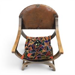 Early 20th century mahogany Savonarola chair, arched leather back with brass studwork, curved open arms with incised geometric decoration, loose patchwork velvet cushion seat, X-frame supports united by circular side rails, on scrolled feet