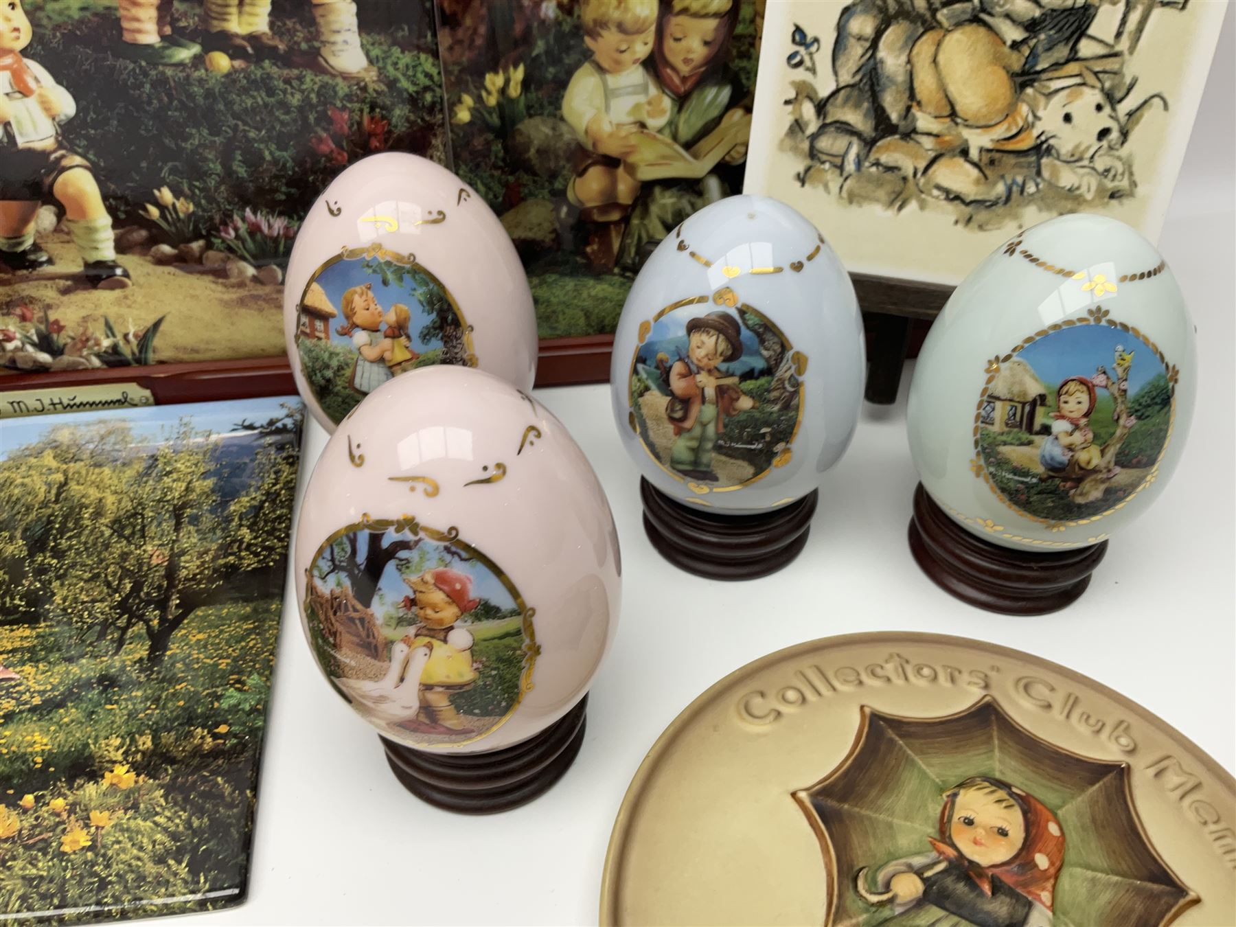 Two ceramic Hummel postcards by Goebel, both displayed on wooden easels, together nine Hummel eggs, set of four Hummel School Days plates, on wooden hanging rack, Goebel Collectors Club Member plaque etc