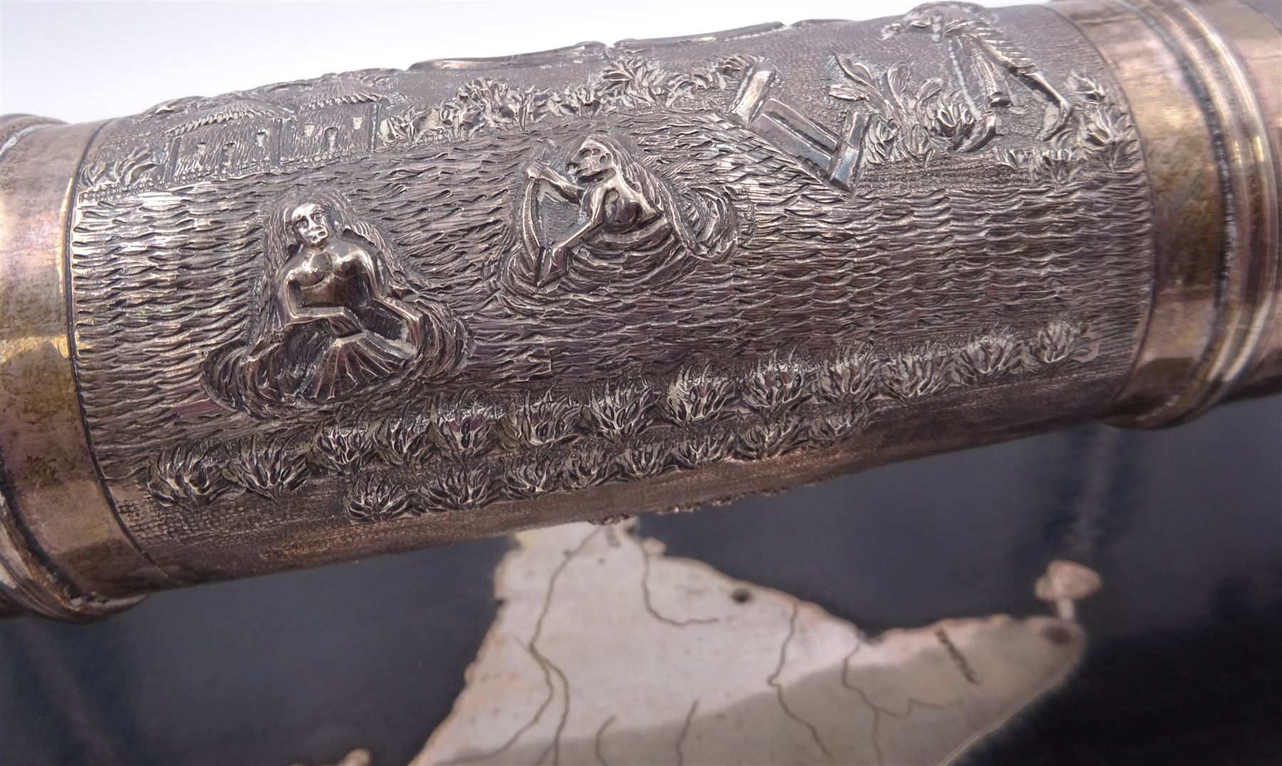 Late 19th/early 20th century Indian silver scroll holder, of cylindrical form with vacant cartouche and embossed with figures, animals and huts, upon a shaped ebonised stand with silver supports and silver panel of India