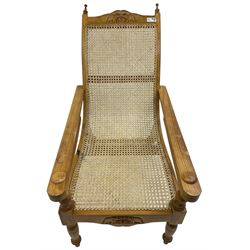 Late 20th century teak framed plantation chair, cresting rail and apron carved with two elephants with upraised trunks, flanked by finials, cane back and seat, fitted with hinged extending leg rests, on ring turned supports