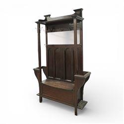Arts and Crafts design oak settle, with projecting moulded cornice over a boarded back fitted with a rectangular bevelled mirror plate and a row of wrought iron coat hooks, the panelled back with arched fielded panels, flanked by shaped umbrella or stick stands with removable drip trays, the plank hinged seat over a panelled box base, on square section supports