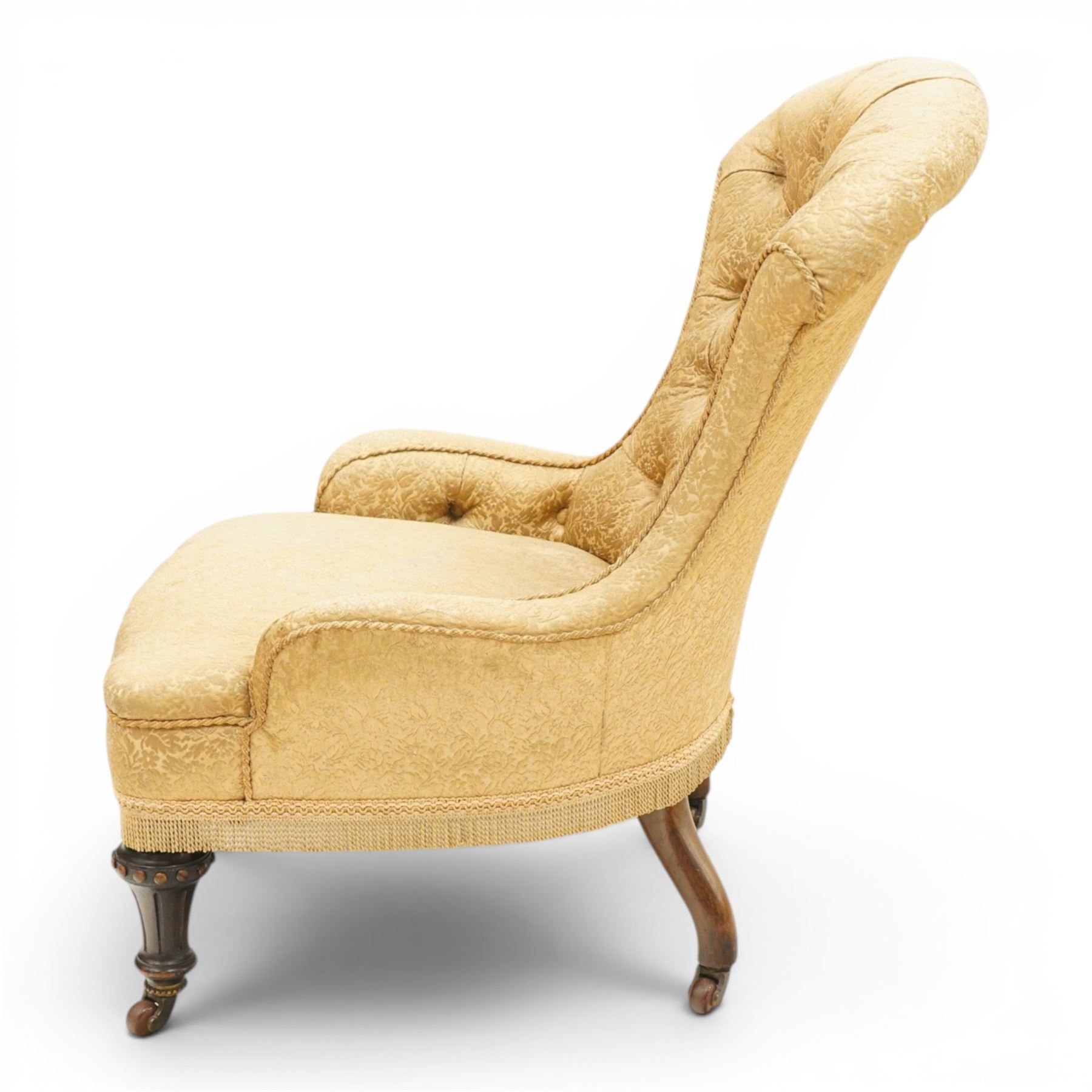 Victorian Aesthetic Movement walnut framed nursing chair, the fan back and sprung seat upholstered in buttoned foliate patterned gold damask fabric with fringing, raised on beaded tapering fluted supports with ceramic castors
