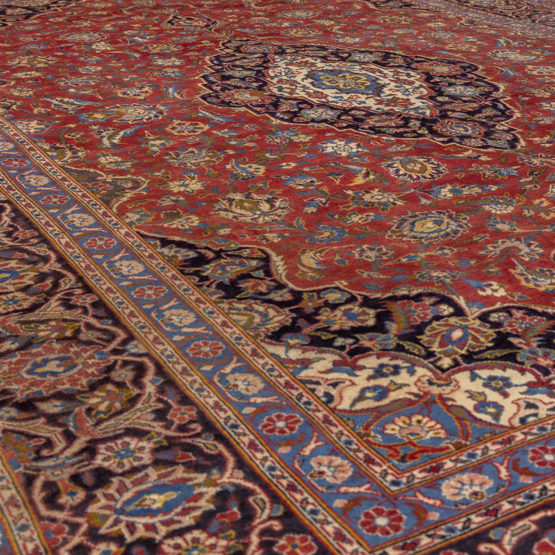 Central Persian Kashan hand-knotted carpet, madder red field with a central cusped medallion of ivory and deep indigo ground, enclosing a stylized floral rosette, framed by scrolling arabesques, field richly ornamented with an intricate network of flowering vinery, palmettes, and delicate floral sprays, spandrels in deep indigo, principal border of navy ground decorated with a continuous palmette and vine-scroll, flanked by multiple guard stripes, with cotton fringe ends 