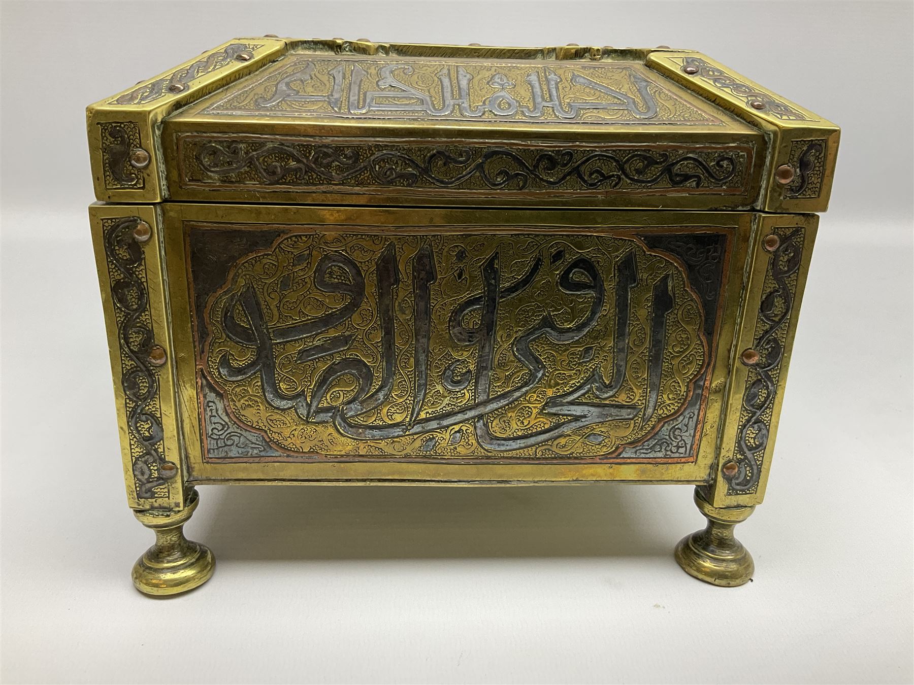 Late 19th century Damascus Cairoware Quran box, of square form with canted hinged cover, upon four circular pad feet, the brass exterior inlaid with silver and copper script and decorative detail, opening to reveal a marquetry lined interior, H15.5cm W19cm D19cm