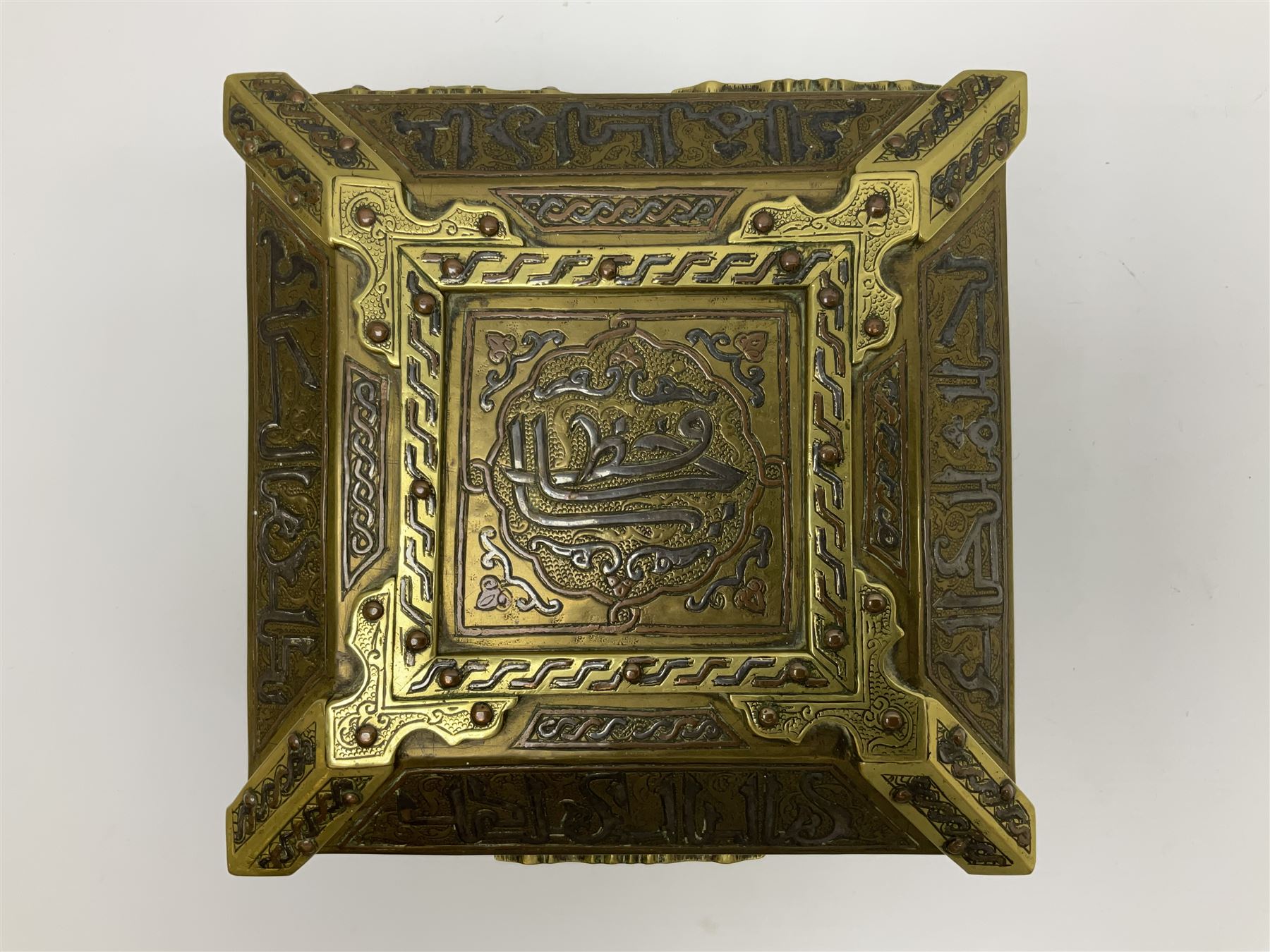 Late 19th century Damascus Cairoware Quran box, of square form with canted hinged cover, upon four circular pad feet, the brass exterior inlaid with silver and copper script and decorative detail, opening to reveal a marquetry lined interior, H15.5cm W19cm D19cm