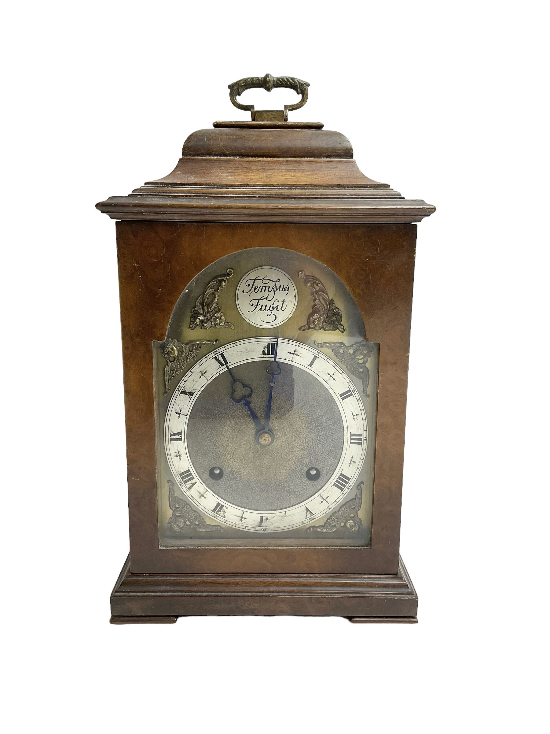 Imperial - English 1950s walnut and mahogany 8-day veneered mantle clock in an18th century-style case, with an inverted bell top and handle, break arch brass dial with a matted centre, silvered chapter ring with roman numerals and trefoil steel hands, cast spandrels and silvered Tempus Fugit boss to the arch, with a rack striking movement striking the hours and half hours on a gong. With pendulum and key.