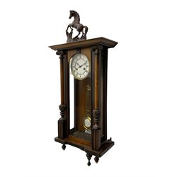 A late 19th century German spring driven 'Vienna' wall clock with a fully glazed door flanked by turned columns with ionic capitals and carving, replacement pediment depicting a prancing horse, with a two-piece enamel dial, Roman numerals, minute track and gothic steel hands, 8-day movement striking the hours and half hours on a gong,  with a gridiron pendulum and beat plate. With key.


