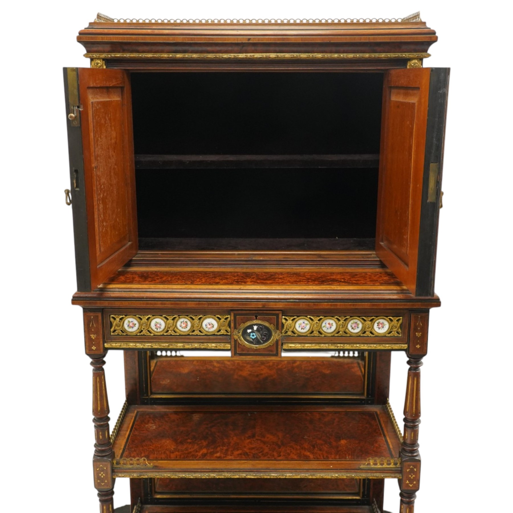 Fine late 19th century Aesthetic Movement ebony and amboyna cabinet on stand with parcel-gilt detail, the moulded cornice with pierced gilt-brass three-quarter gallery above a pair of panelled doors centred by oval porcelain portrait plaques within gilt-metal ribbon-tied frames, the doors further enriched with gilt-metal foliate mounts and flanked by classical figural terms, enclosing a shelved interior, the projecting lower section with amboyna top above a frieze drawer set with a series of floral porcelain roundels within pierced gilt-metal mounts and a central oval floral plaque, raised on turned and fluted supports with gilt-heightened detail, the mirrored back behind two amboyna shelves with pierced gilt-brass galleries, on turned feet