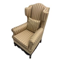 William and Mary style wingback armchair, upholstered in gold striped fabric, on walnut scroll carved supports joined by scroll and foliate carved middle rail, turned H-shaped stretchers, on castors