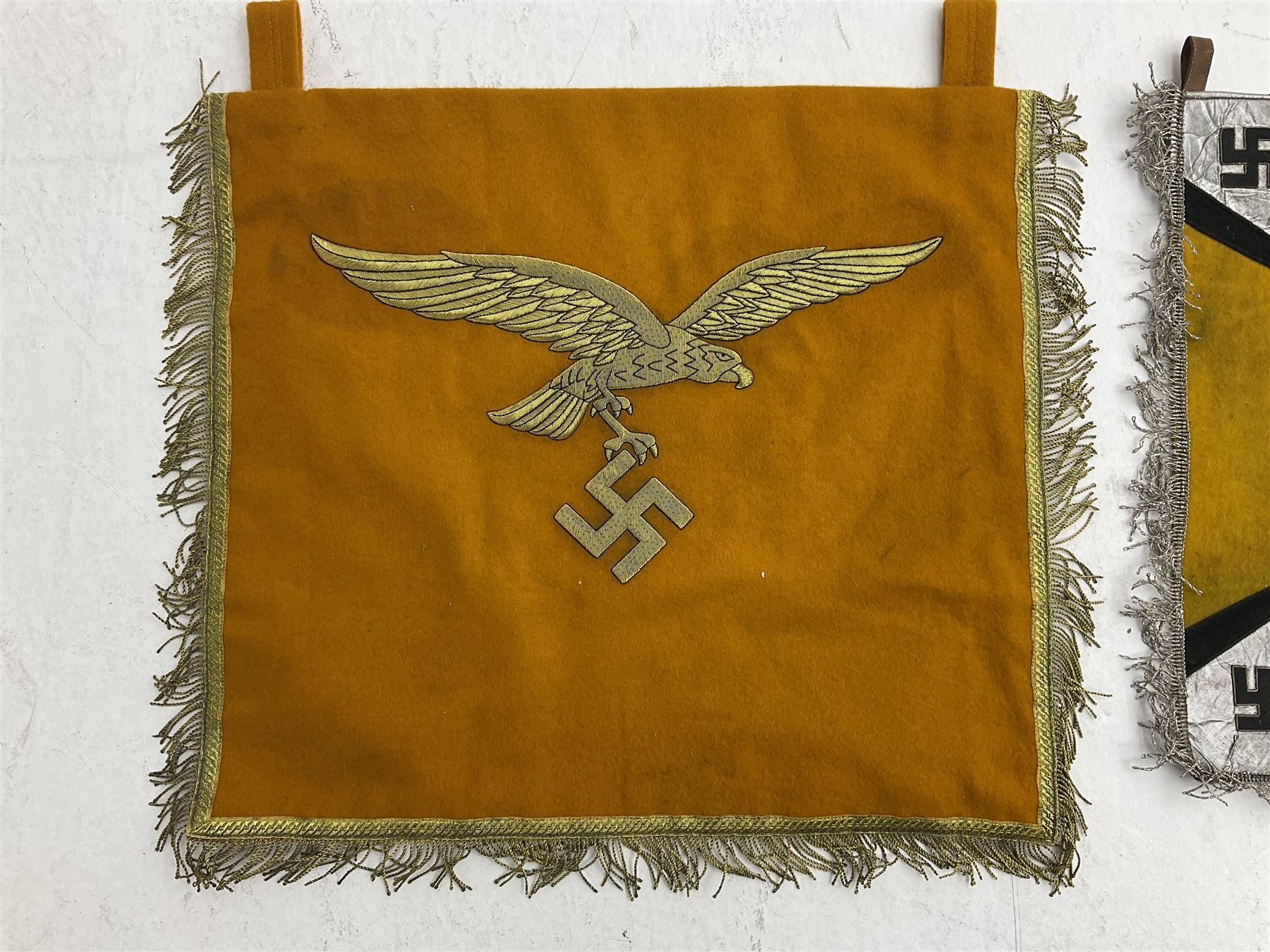 Two replica German Luftwaffe trumpet banners, one heavily silver wire embroidered with Luftwaffe eagle in a wreath, on an amber, white and black rayed background, , with silver wire tasselled border and four leather hanging loops, approx. 50 x 49cm; the other slightly larger heavily gold wire embroidered with the Luftwaffe eagle on a plain amber ground with gold wire tasselled border and two hanging loops (2)