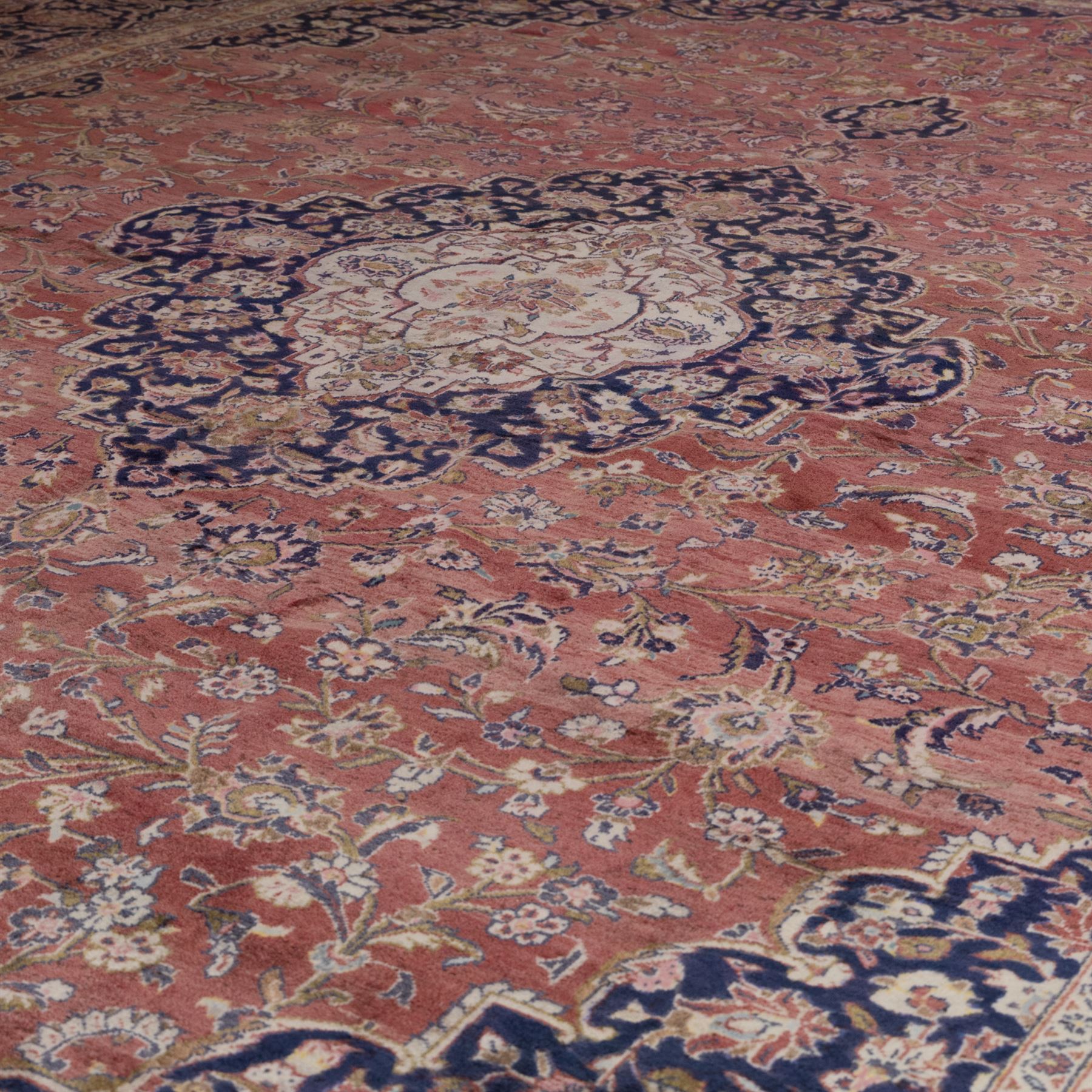 Large Central Persian Kashan hand-knotted carpet, madder red field with central cusped medallion on beige ground enclosed by indigo border with an intricate all-over arrangement of scrolling vines and floral motifs within conforming spandrels and a indigo ground main border ornamented with floral designs, enclosed by guard stripes and cotton fringe ends 