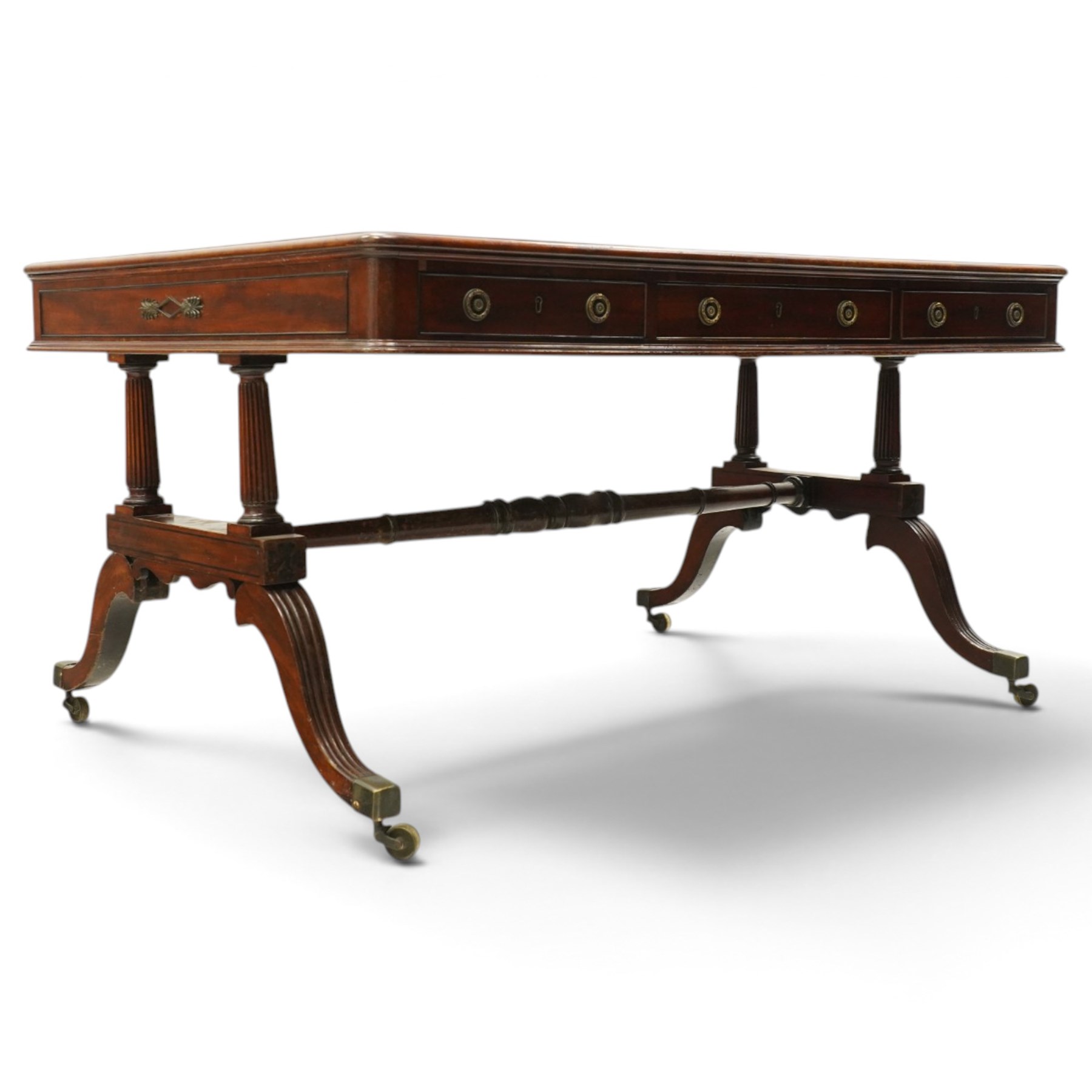 Regency inlaid mahogany and brass mounted library table, the rectangular top inset with gilt tooled burgundy leather writing surface, enclosed by  crossbanding and boxwood stringing with roundel corner motifs, above three cock-beaded frieze drawers to either side fitted with brass ring handles and brass escutcheons, the end friezes centred by applied brass foliate mounts, raised on twin reeded column supports over rectangular platform ends, united by a ring-turned stretcher, the reeded sabre legs terminating in brass caps and castors, one drawer bearing ivorine label 'Spillman & Co., St Martins Lane, London'
