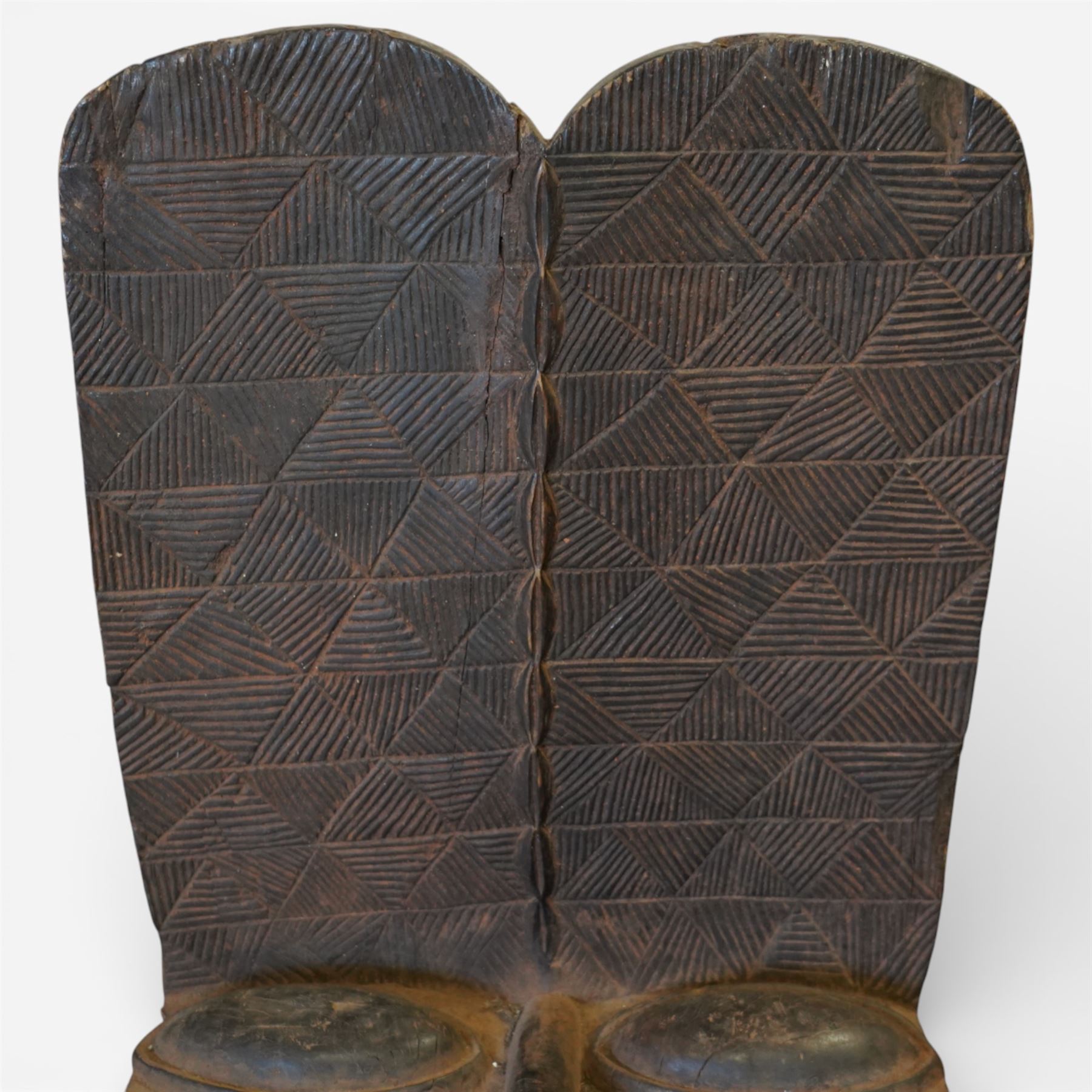 African carved hardwood ceremonial seat, monoxylous construction with twin dished seat wells, the tall back incised with repeating geometric triangular motifs, the front carved in the form of a tribal mask with projecting features, raised on a cylindrical base with ribbed and textured carving
