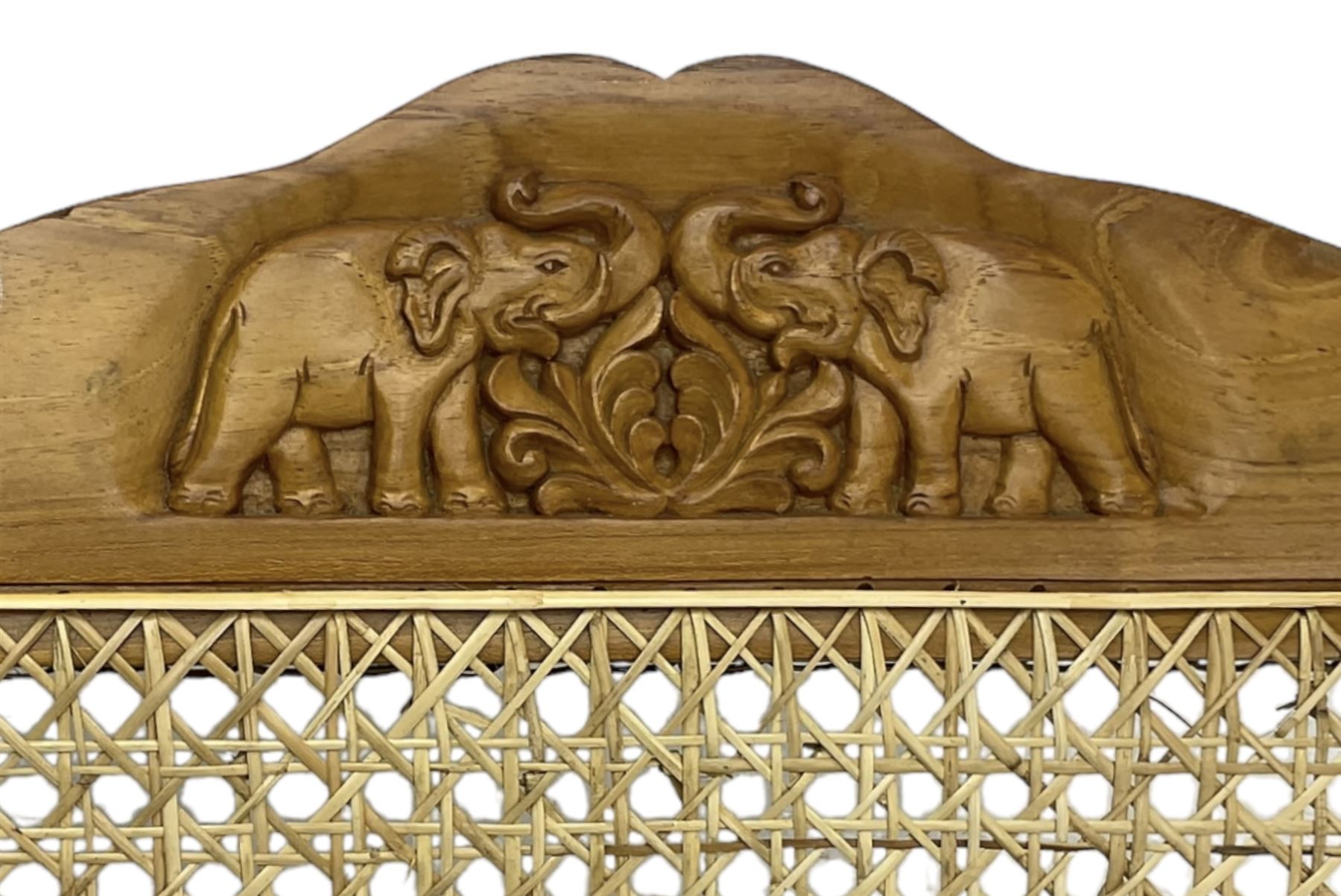 Late 20th century teak framed plantation chair, cresting rail and apron carved with two elephants with upraised trunks, flanked by finials, cane back and seat, fitted with hinged extending leg rests, on ring turned supports