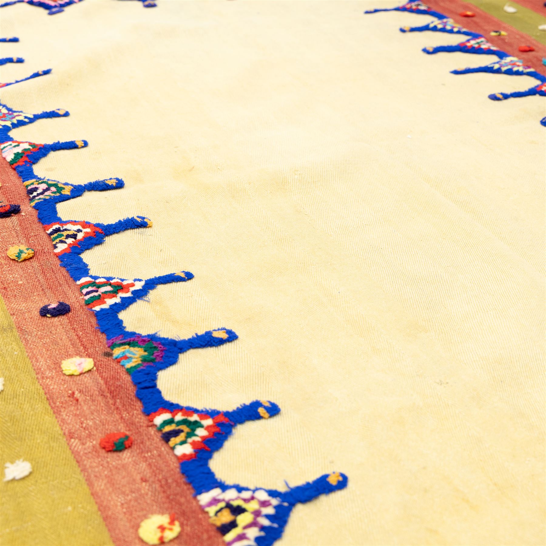 Persian Jajim flatweave kilim, plain beige central field, framed by stepped indigo and polychrome inner border within broad striped outer bands in rose, green, and ivory, decorated with knotted wool tufts and fringe ends 