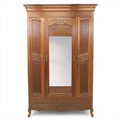 Early 20th century French oak triple door armoire, the moulded top above a frieze with carved lozenge decoration and central rosette, the central mirrored door enclosed by carved surround with floral crest, flanked by panelled doors with lattice carving, the base with shaped apron centred by carved basket motif, upon shaped cabriole supports