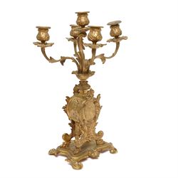 French-Late 19th century 8-day gilt brass rococo style mantel clock, c1890, waisted case  decorated with scroll work, leaves and musical instruments, resting on an attached plinth with splayed feet, dial within a glazed brass bezel with a gilt repoussé centre, porcelain cartouche Roman numerals and steel fleur-di-Lis hands, with a twin train movement, recoil anchor escapement and countwheel strike, sounding the hours and half-hours on a bell. With a matching pair of five-light candelabra. No Pendulum or Key.