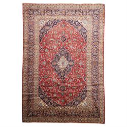 Persian Kashan hand-knotted carpet, madder red ground centred by shaped dark navy and ivor...
