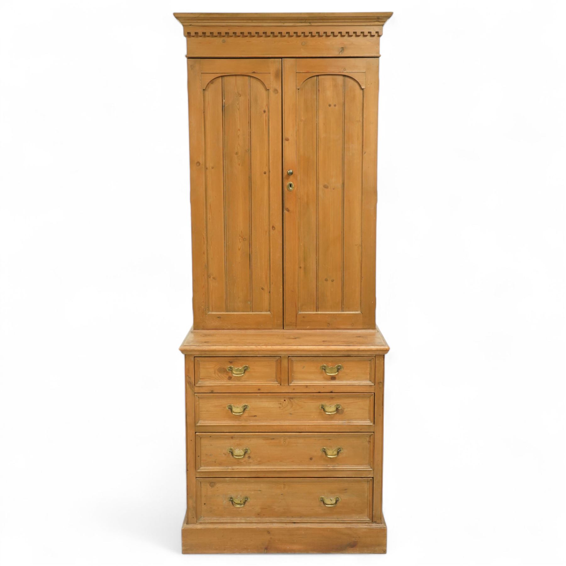 Early 20th-century pine gun cabinet with later fitted shelves, of two-part construction, upper cupboard moulded and dentil cornice above two panelled doors revealing three removable shelves and circular recesses for gun barrels at the top, lower section with two short and three long graduated drawers with brass swan neck handles on plinth base 