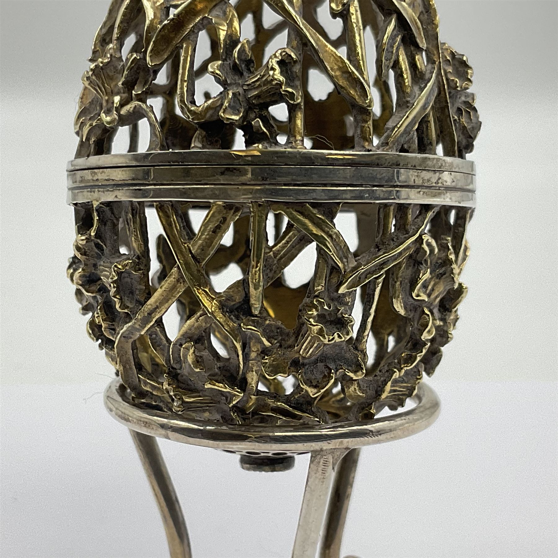 Modern silver limited edition Easter egg, no. 70/500, the gilt openwork body decorated with daffodils and detachable cover with pierced circular panel set with a single faceted blue stone, opening to reveal a gilt interior, upon silver stand with three scrolling pad feet, each hallmarked St James House Company, London 1980, height including stand 8cm