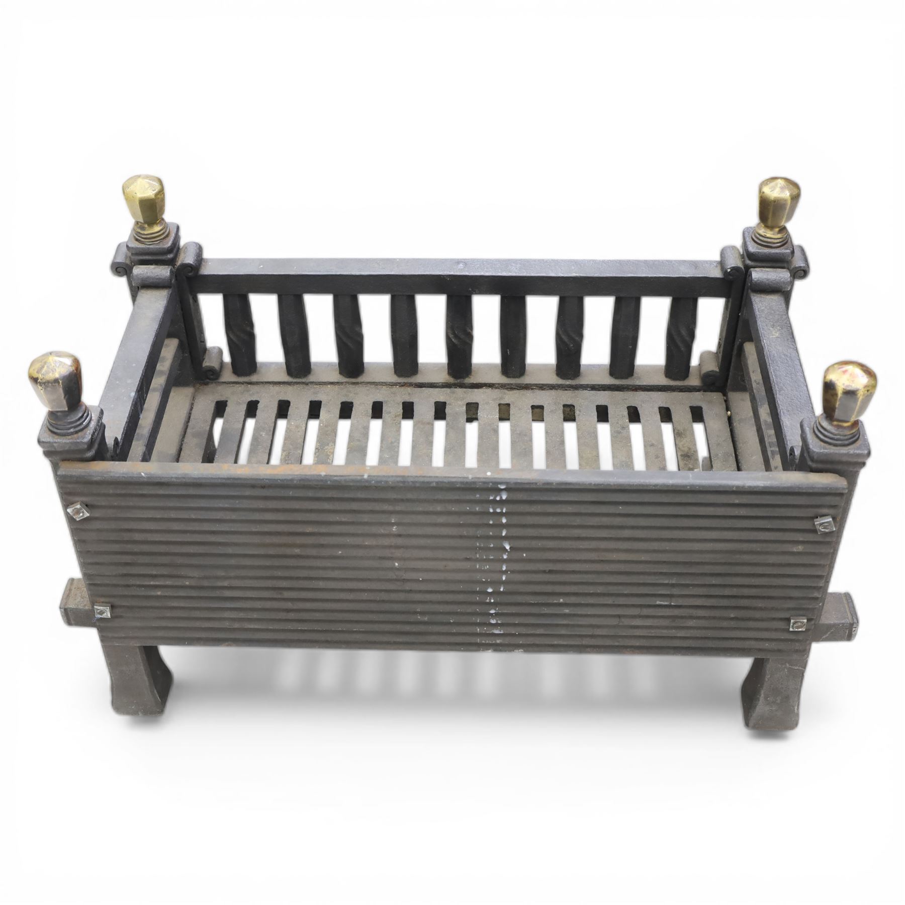 Arts and Crafts cast iron fire grate, rectangular backplate with plain upper rail over a slatted front of cylindrical tapering bars, flanked by square section uprights with fluted detail and scroll terminals, raised on stepped feet with shaped brass roundel mounts and brass finials to the corner posts