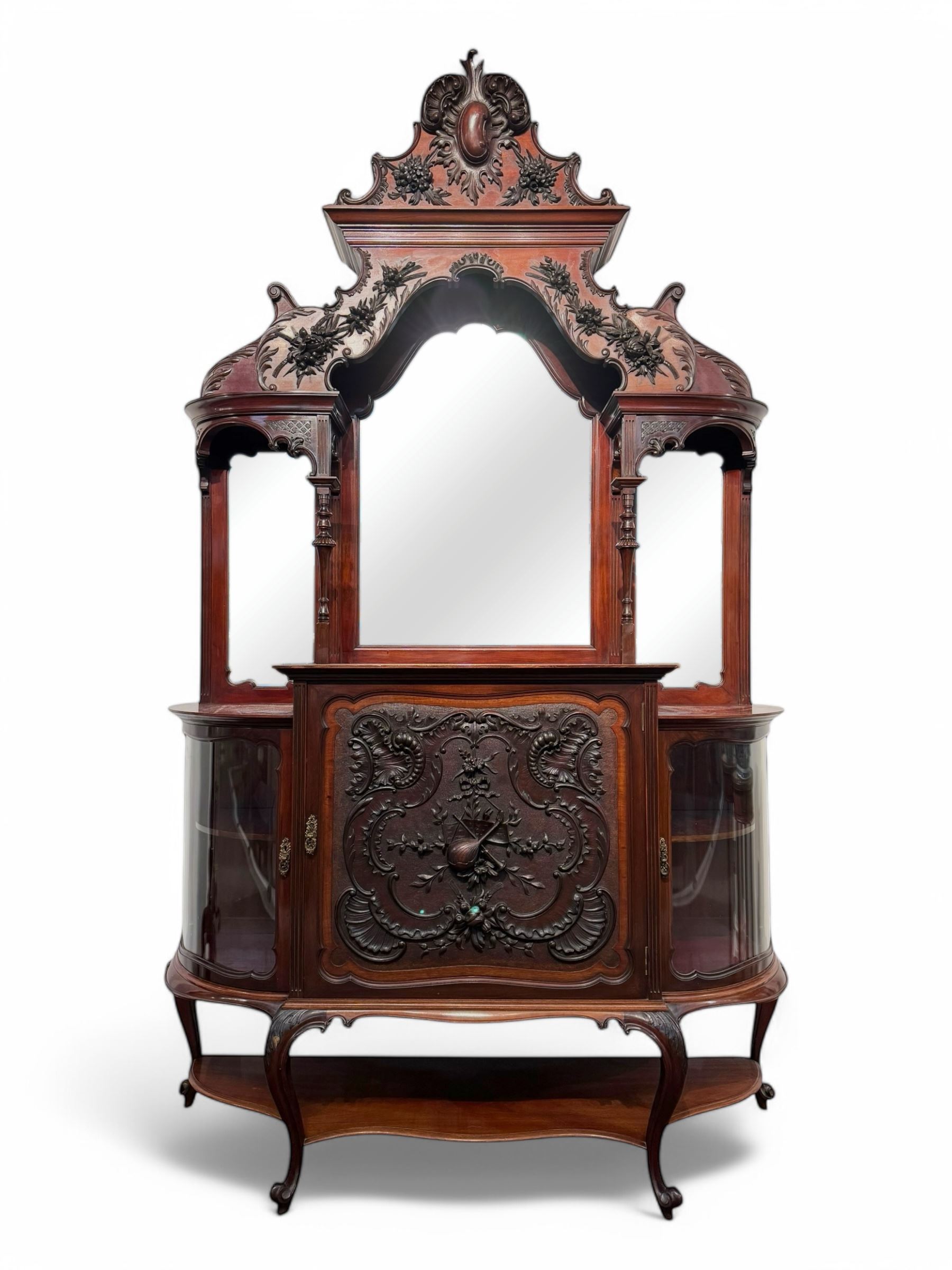 Late 19th century mahogany mirror back cabinet, shaped C-scroll pediment decorated with carved acanthus leaf cartouche and shells, shaped canopy top with applied foliate carvings over triple bevelled mirror back, central cupboard enclosed by panelled door carved with musical instruments, scrolls and shells, flanked by two glazed cabinets, on acanthus leaf carved cabriole supports united by undertier 