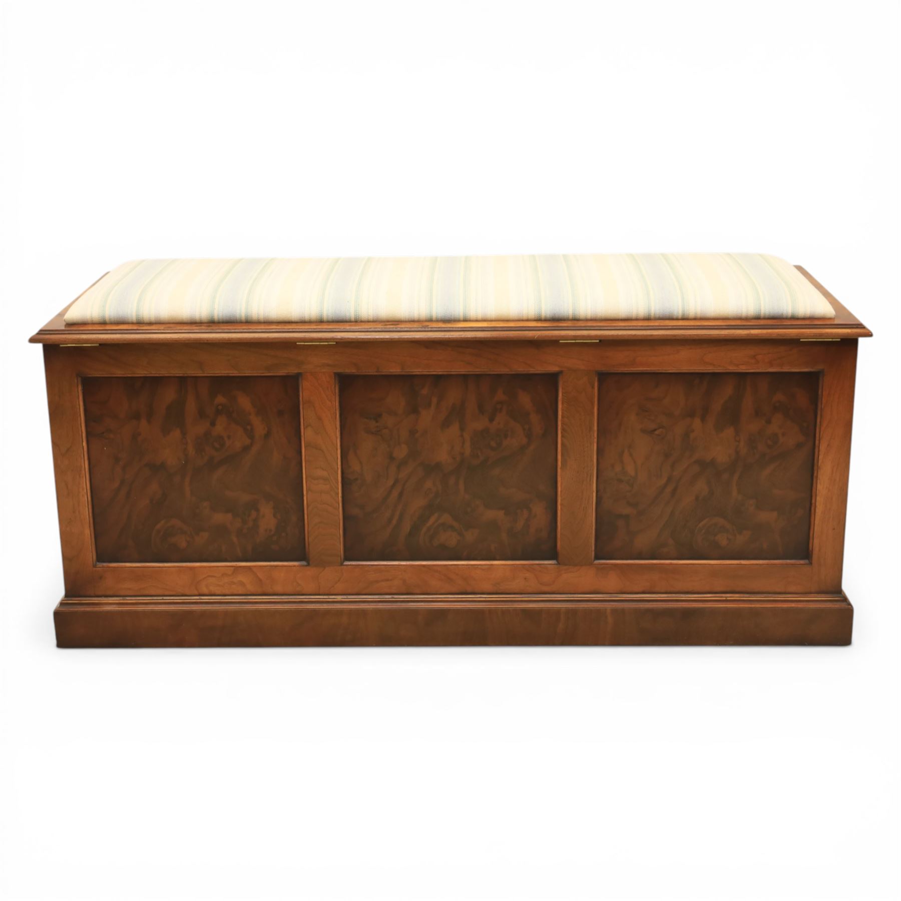 Traditional design figured elm blanket chest, with padded striped upholstered seat, over a hinged top with panelled sections below raised on a plinth base