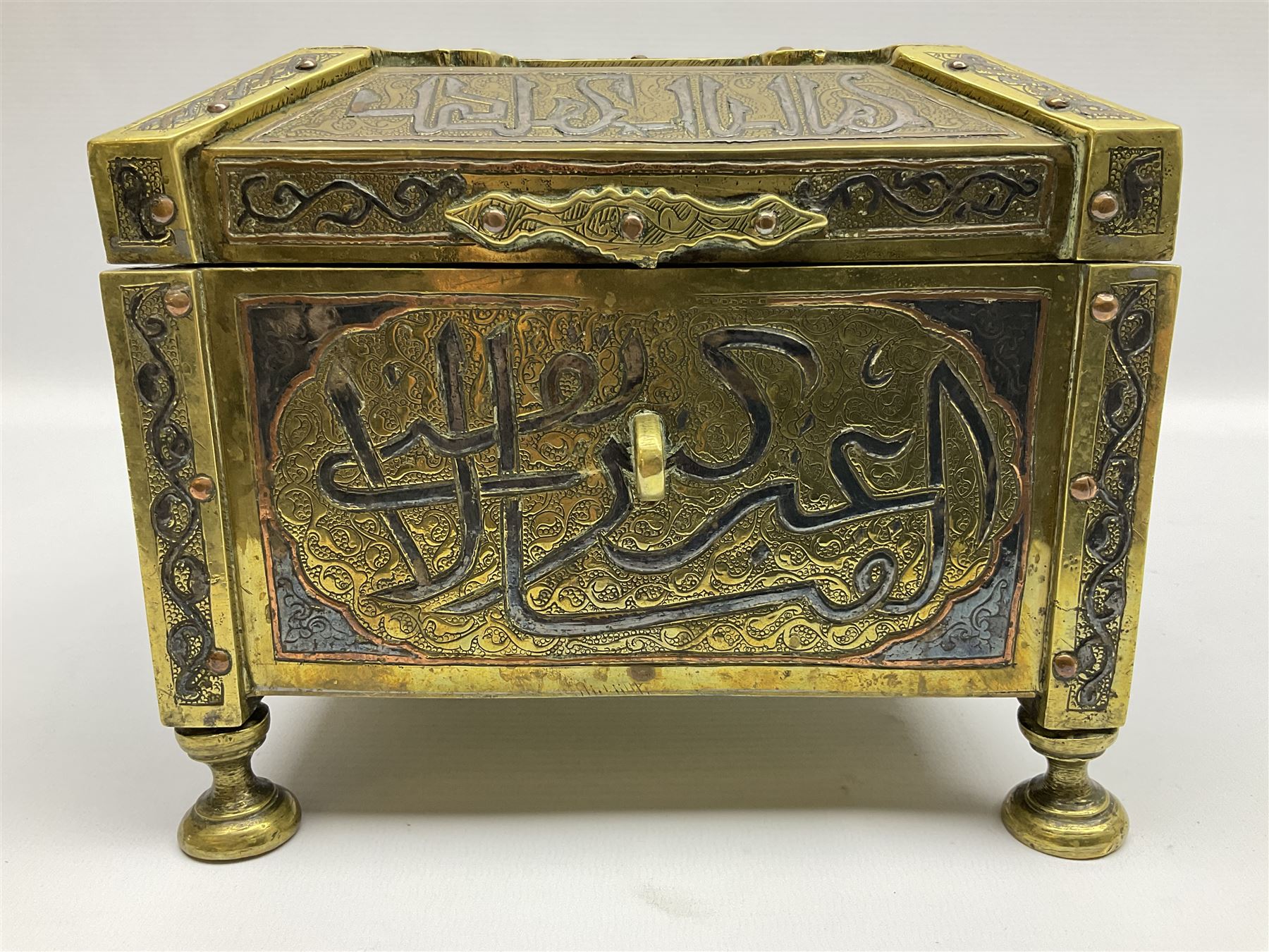 Late 19th century Damascus Cairoware Quran box, of square form with canted hinged cover, upon four circular pad feet, the brass exterior inlaid with silver and copper script and decorative detail, opening to reveal a marquetry lined interior, H15.5cm W19cm D19cm