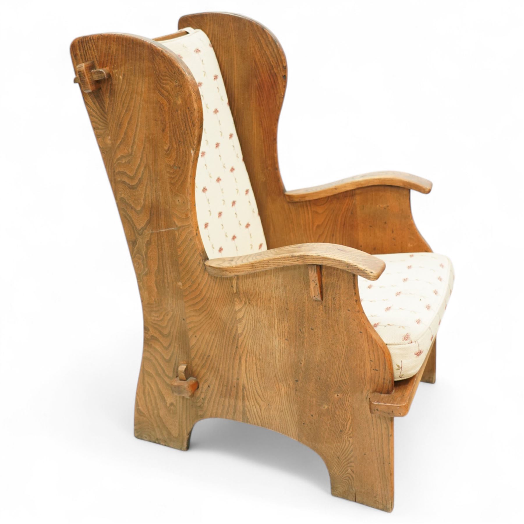 20th century pair of country lambing chairs in light oak, each with high winged back and shaped arms, pegged construction with panelled sides and arched aprons, loose seat and back cushions upholstered in cream floral fabric