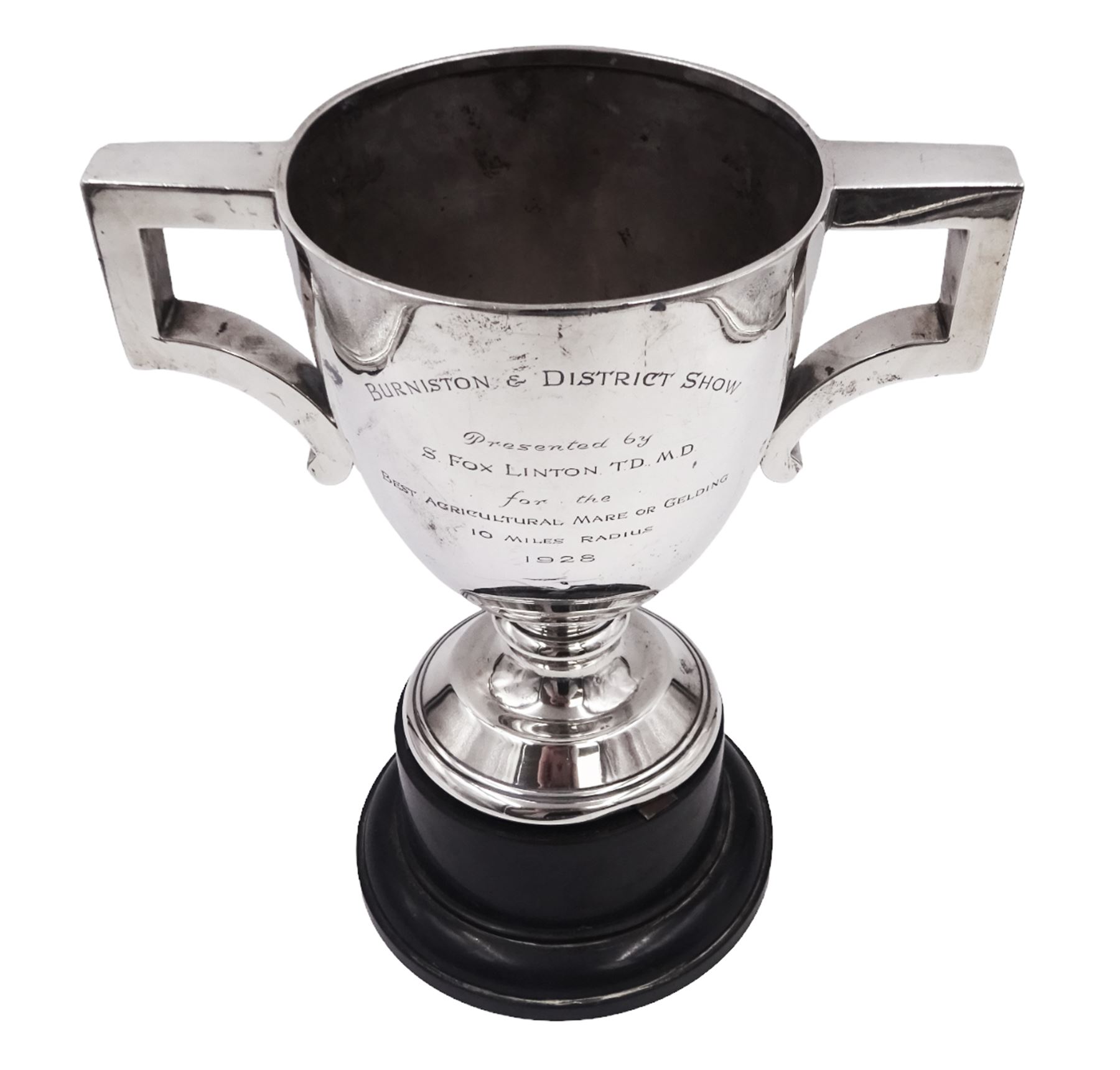 1920s silver trophy cup, with angular twin handles, upon knopped stem with circular spreading foot, the body with presentation engraving 'Burniston & District Show, presented by S Fox Linton TD MD, for the Best Agricultural Mare or Gelding, 10 miles radius, 1928' and engraved with winners verso, hallmarked Charles Boyton & Son 1921, upon black plastic base, including base 25cm