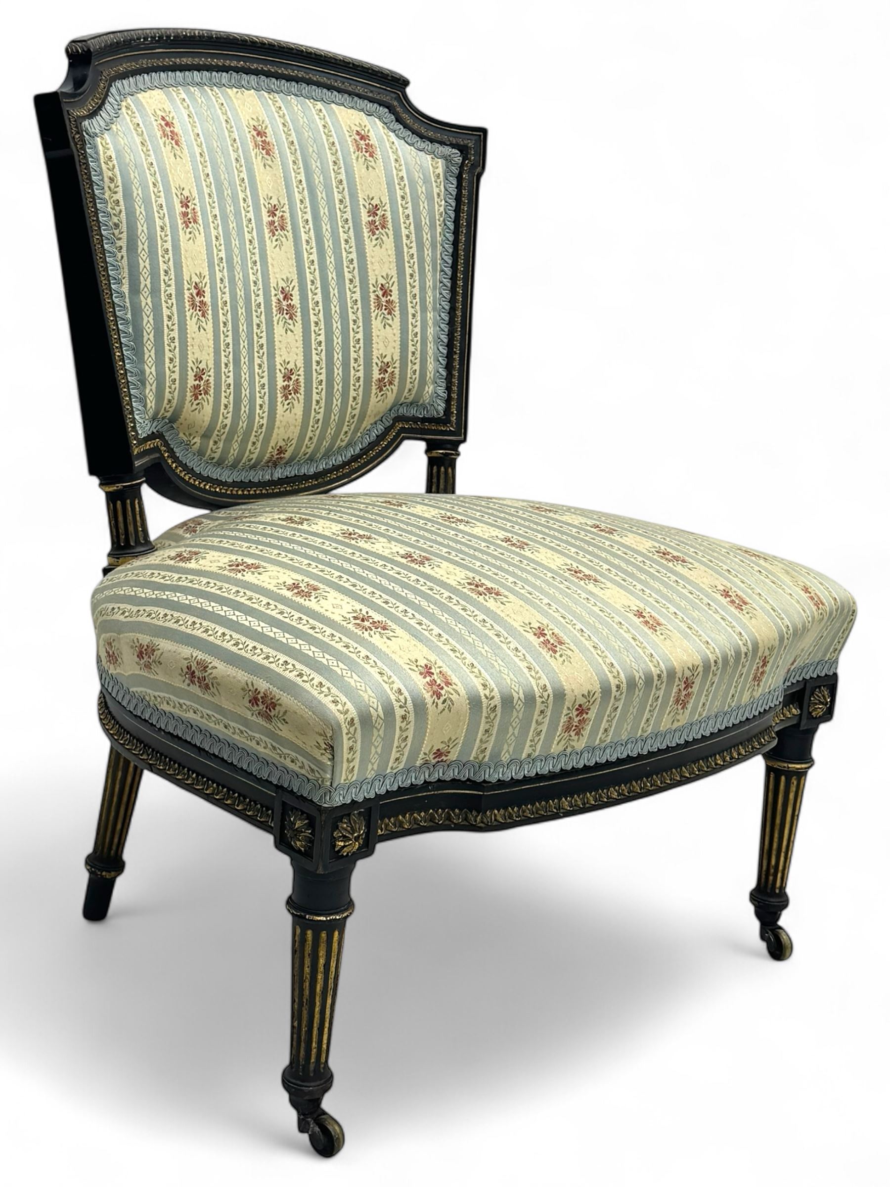 19th century French ebonised low chair, upholstered in floral striped fabric within a moulded frame with gilt line and foliate decoration, the seat of serpentine form, raised on turned fluted legs with toupie feet