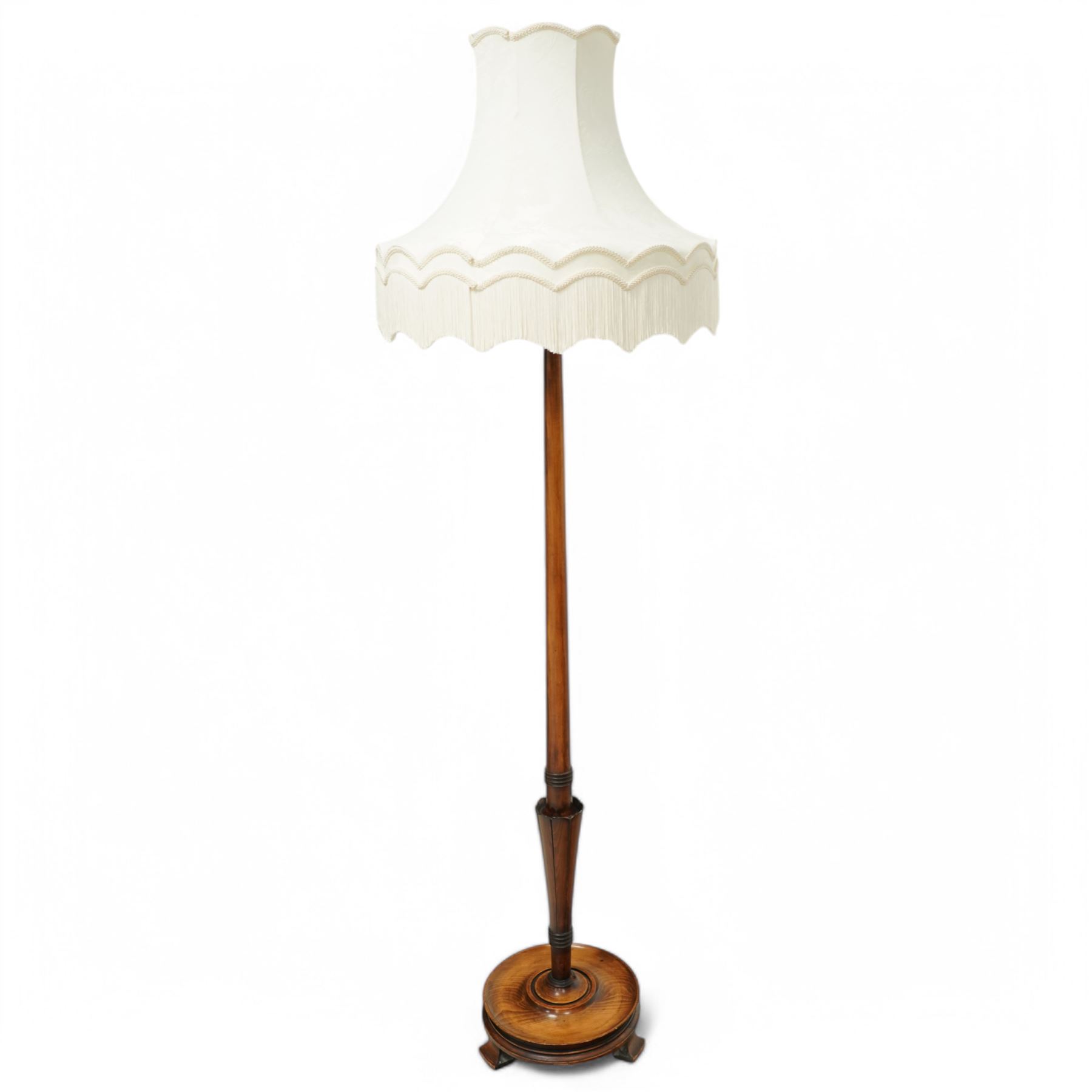 20th century walnut standard lamp, flared and faceted column over circular stepped base with shaped feet, with later scalloped cream fabric shade and fringe trim