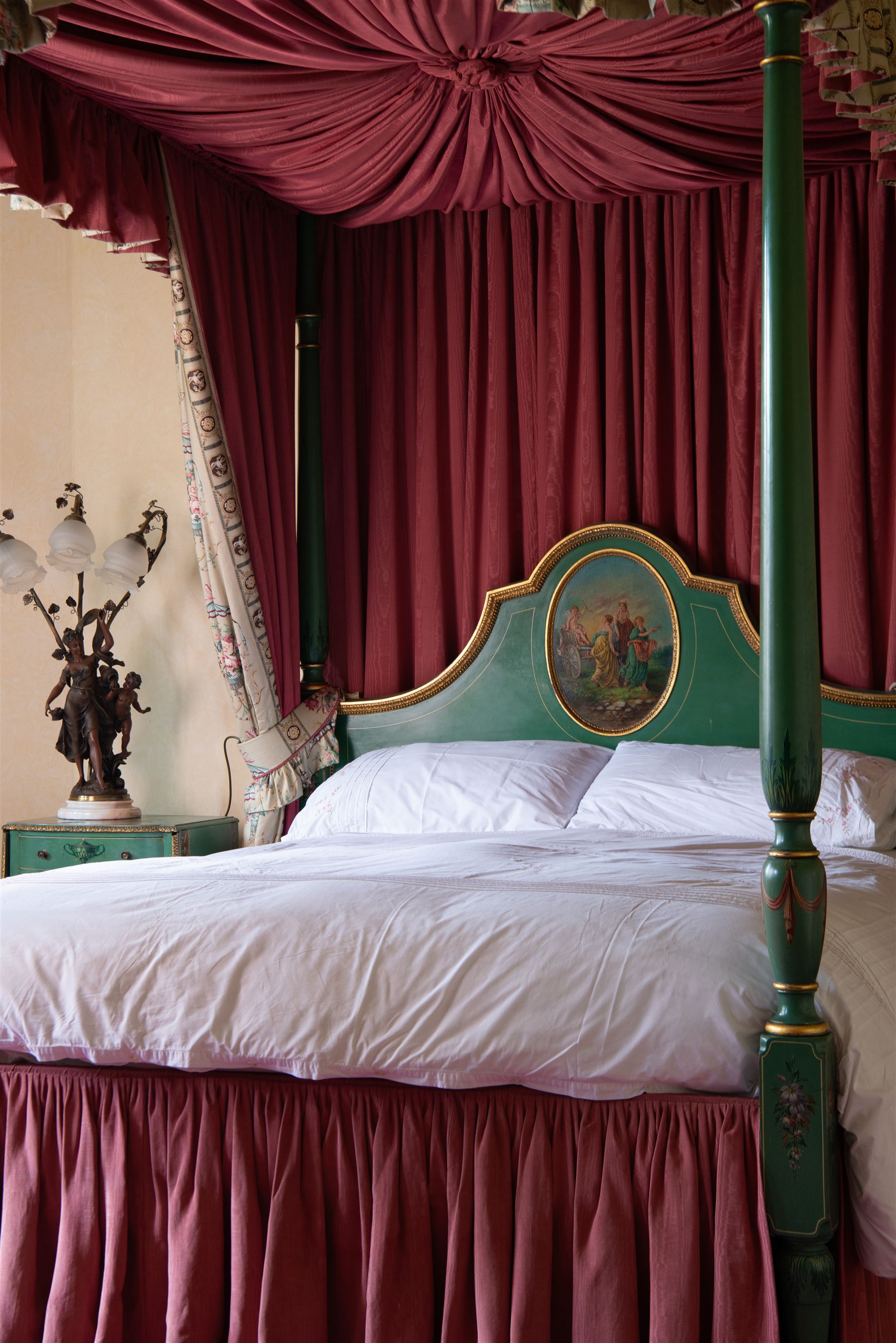 Edwardian Adam Revival green-painted and parcel gilt 5' Kingsize four poster-bed, acanthus leaf moulded canopy with curled feather finials, on four turned posts painted with acanthus leaves and draped festoons, shaped arch headboard with painted oval panel depicting maidens of love leading a chariot holding Eros, with craquelure finish, dressed in silk curtains decorated with floral bouquets 