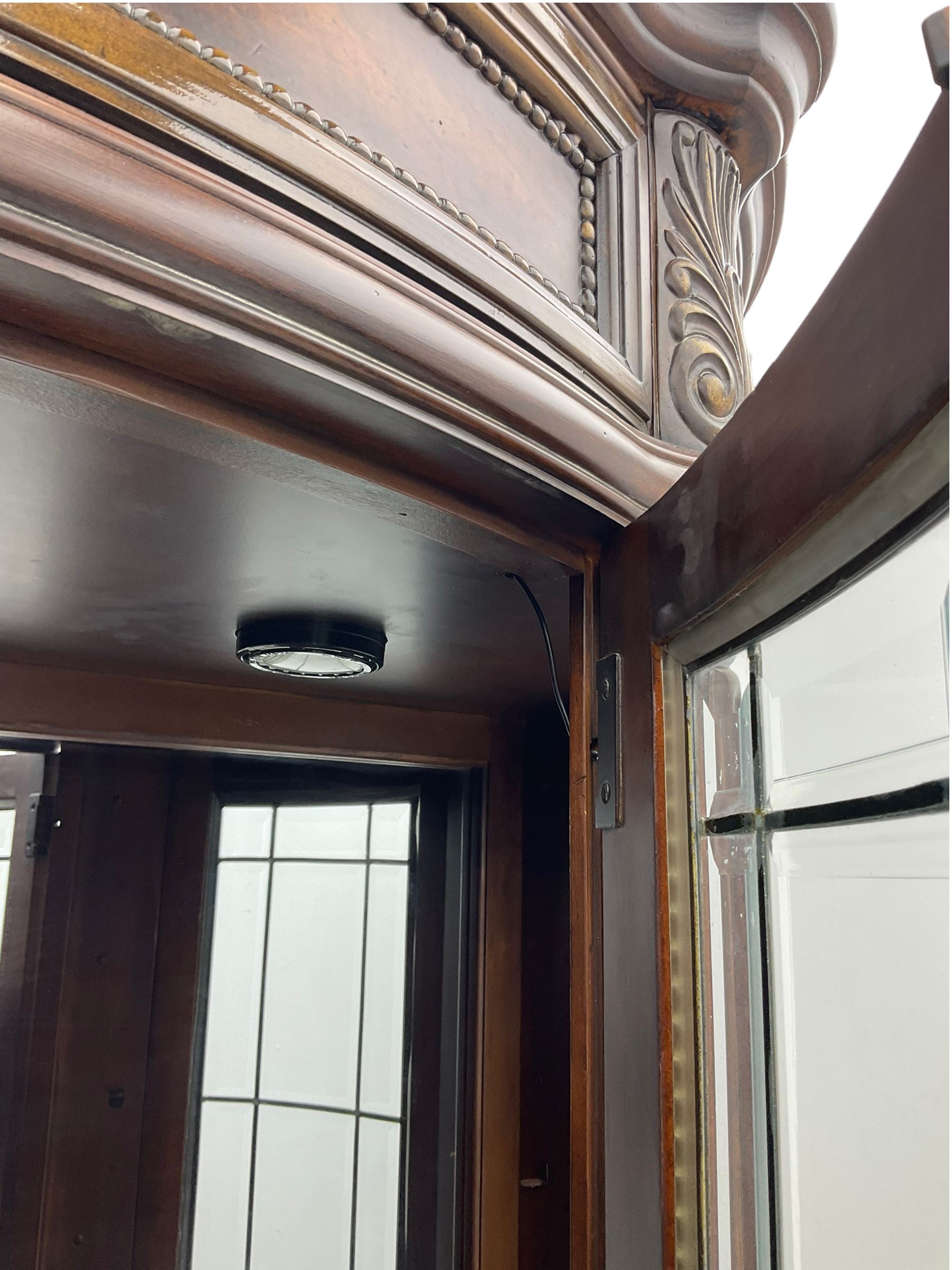  Kevin Charles American walnut demi lune display cabinet with illuminated interior, projecting cornice, two bevel edge glazed doors enclosing two adjustable shelves above single drawer and four cupboard doors with wine rack, carved scrolling supports