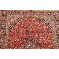 Persian Kashan hand-knotted carpet, madder red ground centred by shaped dark navy, ivory and pale blue floral medallion with pendants, surrounded by scrolling vines, palmettes and flowerheads, dark navy spandrels, enclosed by dark navy floral border with multiple floral guard stripes