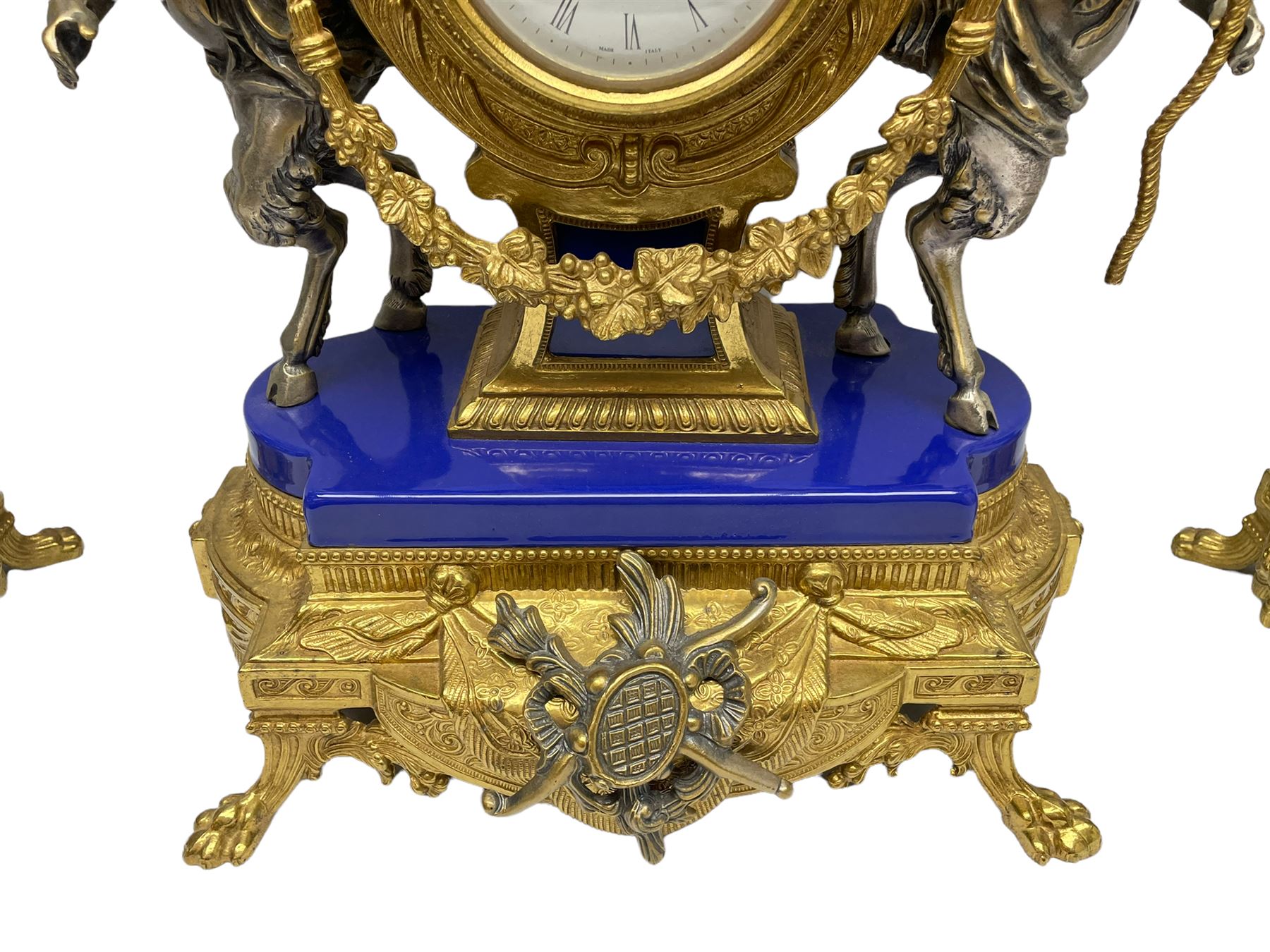 A 20th century continental gilt metal Lyre mantle clock with a pair of matching six light candelabra, spring driven movement housed in a gilt drum case on a brass and cobalt blue base with paw feet, two bronzed metal figures of mythological cherubs with animal legs and cloven hooves supporting a festooned garland swag, eight-day twin barrel striking movement with a floating lever balance escapement, striking the hours and half-hours on two bells, white enamel dial with roman numerals, minute track and pierced steel hands, dial inscribed “Imperial”. 