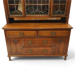 Arts and Crafts oak dresser, projecting moulded cornice above three glazed panels with coloured leaded glass floral panels, flanked by two glazed doors enclosing two adjustable shelves, open mirrored recess below with mirrored back, the base fitted with two short drawers over two central drawers flanked by carved panelled cupboard doors, ornate brass handles, raised on tapering square legs
