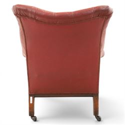Early 20th century style oxblood red leather Chesterfield armchair, deep buttoned shaped back over padded scroll arms and loose seat cushion, raised on turned mahogany front supports with brass caps and castors
