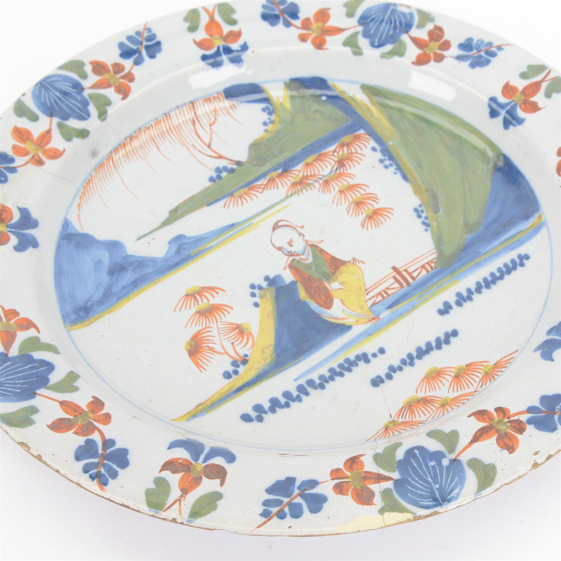 Mid 18th century Lambeth Delft plate, probably from the Abigail Griffith factory, circa 1750, painted in polychrome enamels with a figure seated in a landscape, within a garlanded border of flowers to rim, D34cm, together with a Liverpool Delft plate, circa 1760, painted in blue with a boathouse surrounded by trees by a river, within diaper and cell borders, D37cm